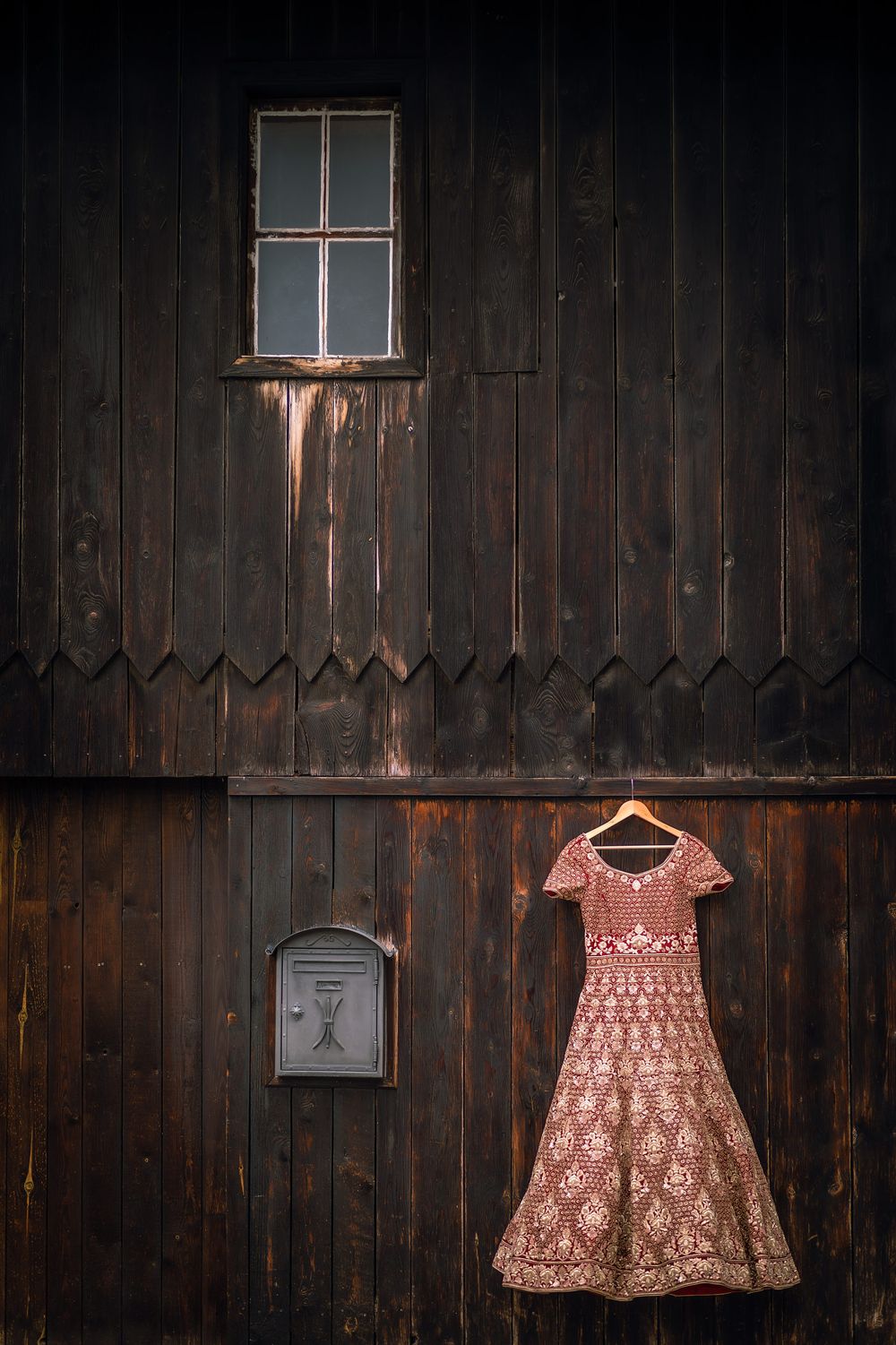 Red embroidered lehenga dress hanging on rustic wooden wall with small window.