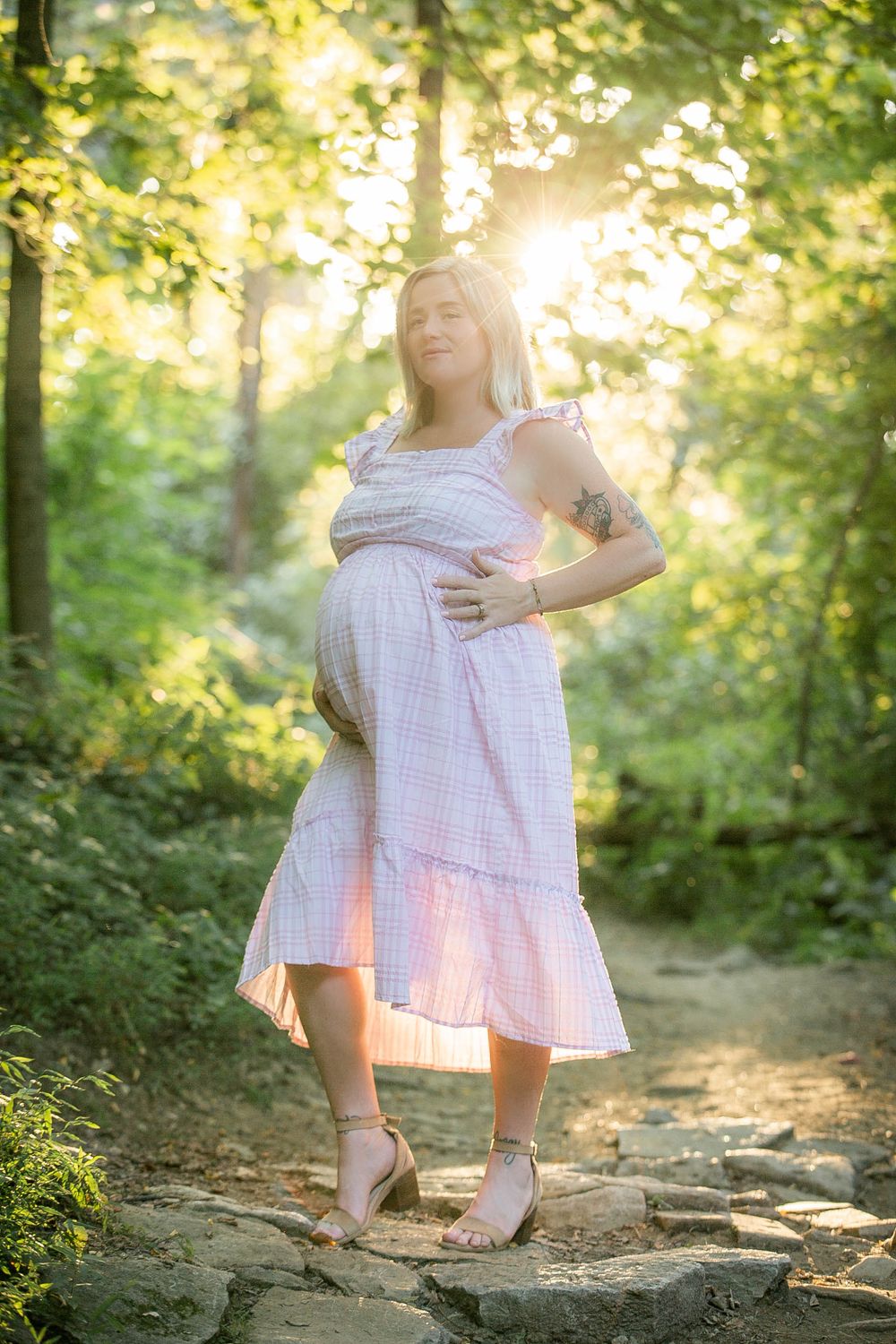Maternity portrait in pink dress on stone path with sunlit forest background.