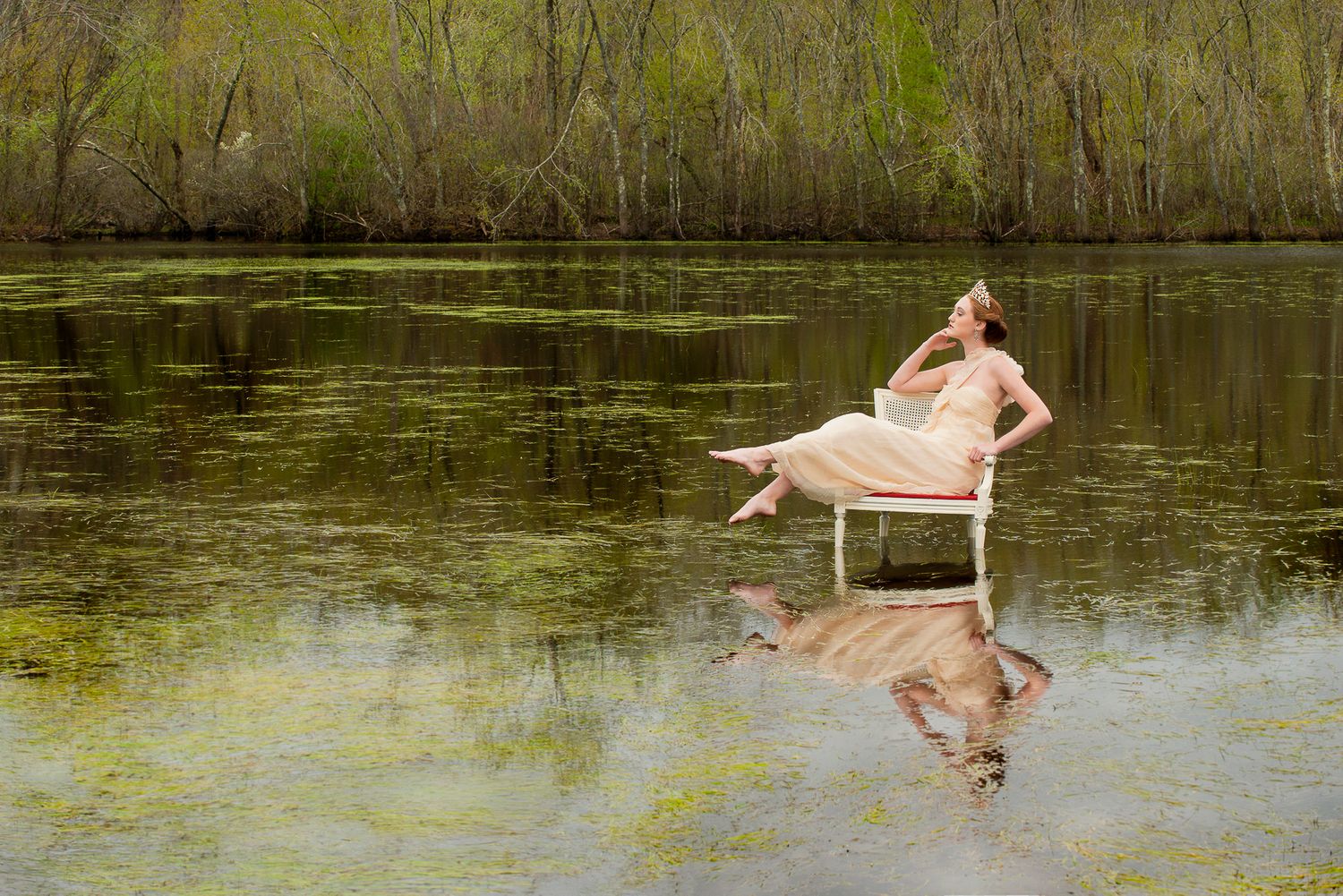 Figure relaxing on white chair submerged in still pond water with reflection.
