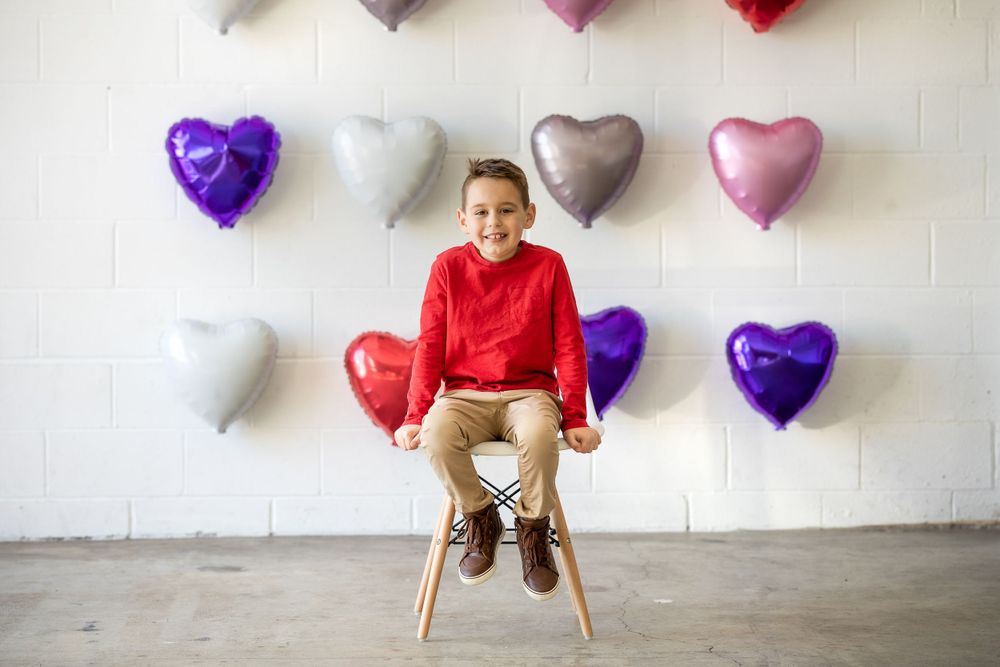 Colorful heart-shaped balloons decorate a white wall above a wooden stool.