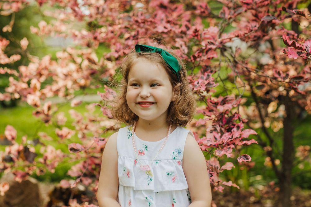 Portrait of a child in white dress with green headband amidst pink cherry blossoms.