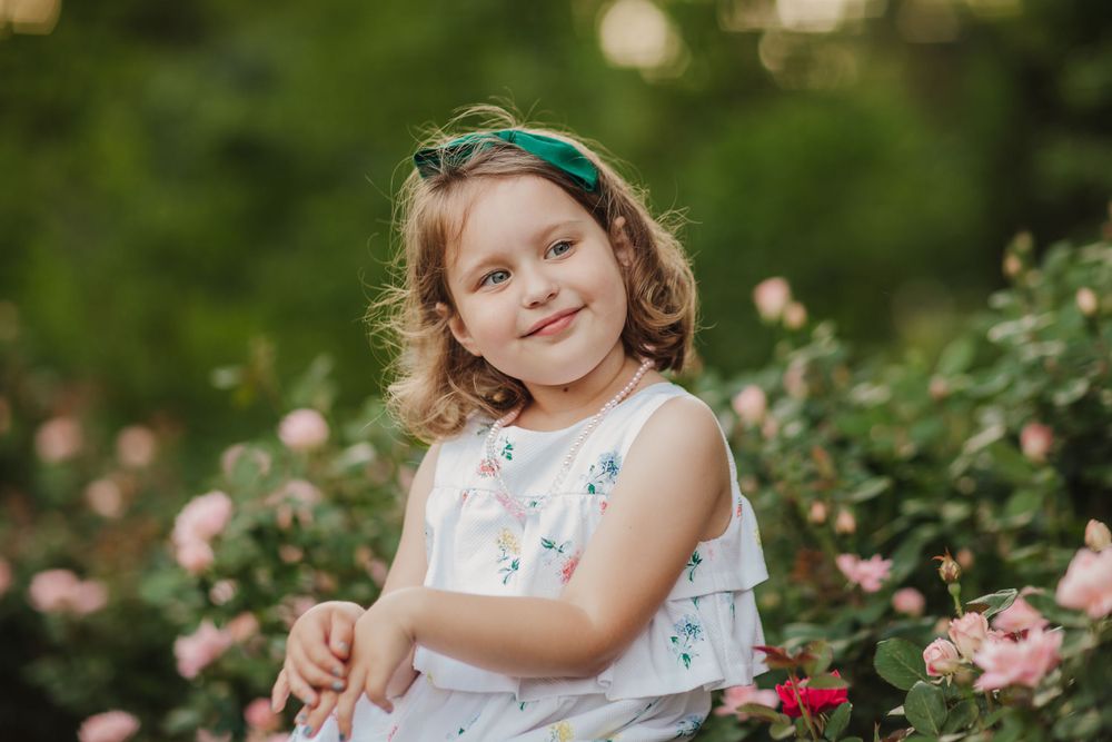 Child in floral dress poses in garden surrounded by pink roses.