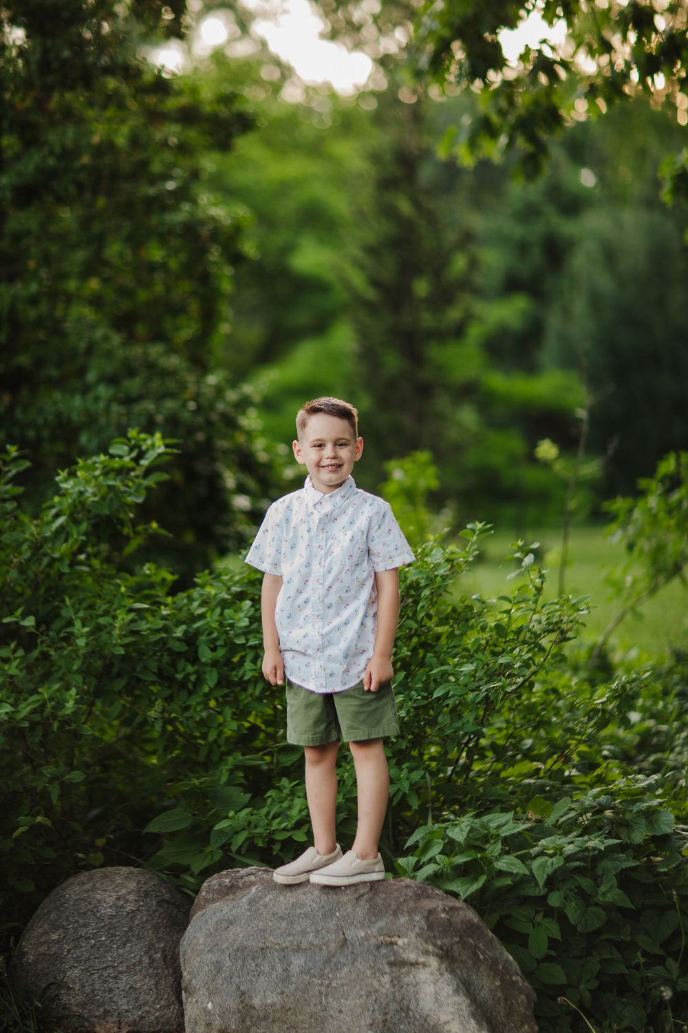Child in white shirt and shorts stands confidently on large rock in lush garden setting.