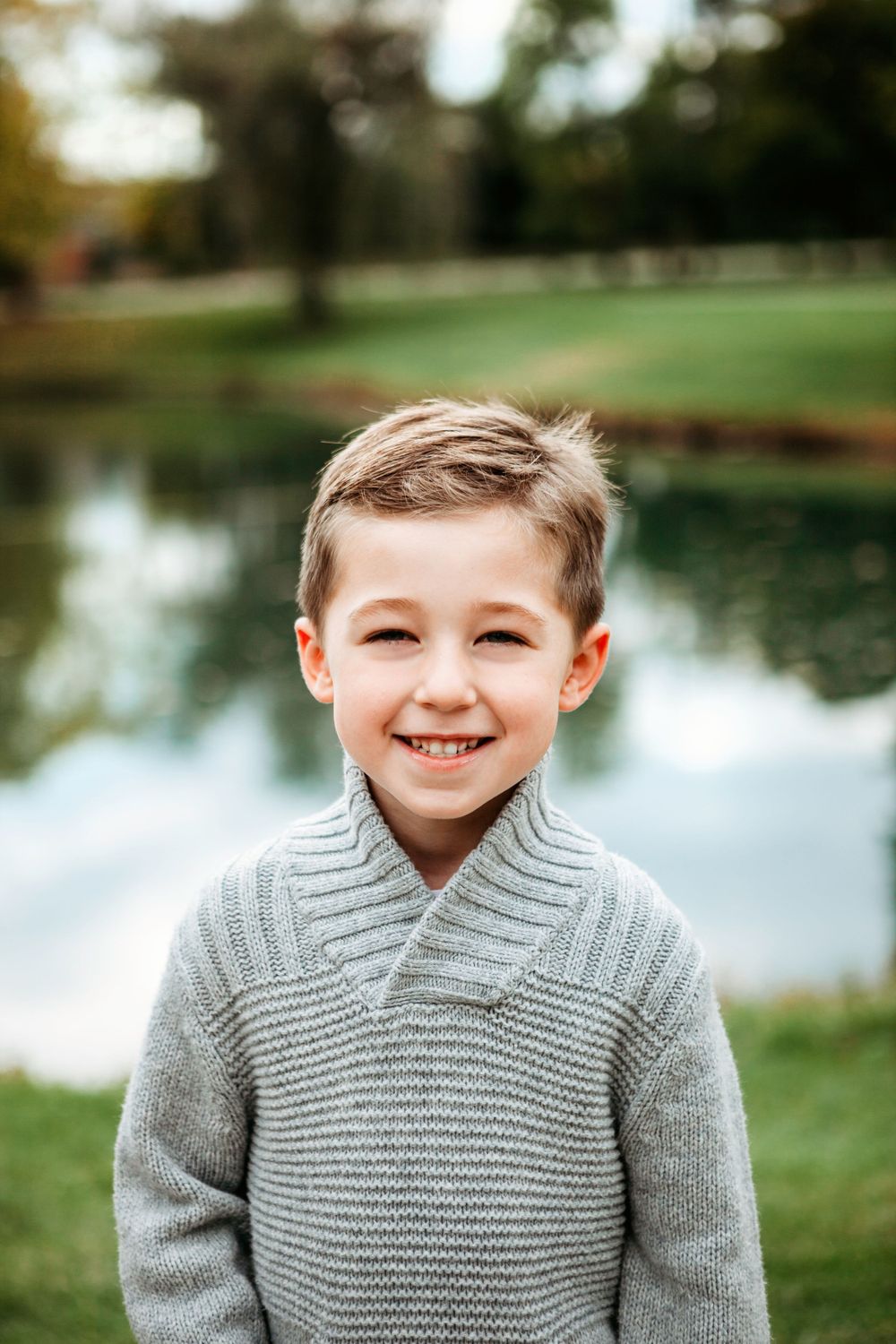 Young child in gray sweater smiling by a pond on a sunny autumn day.
