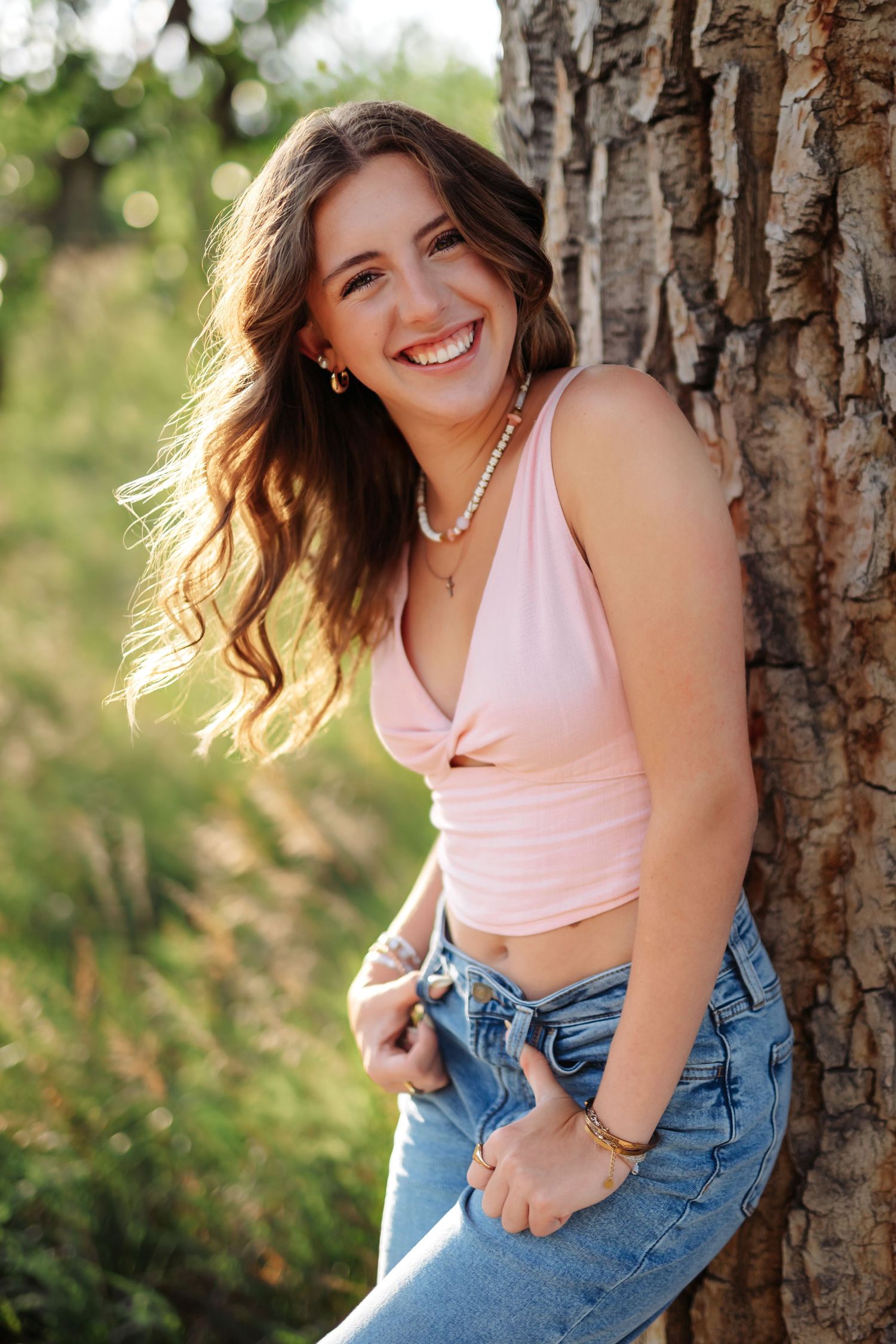 Woman in pink top and jeans poses casually against tree trunk in outdoor portrait session in Navarre Florida