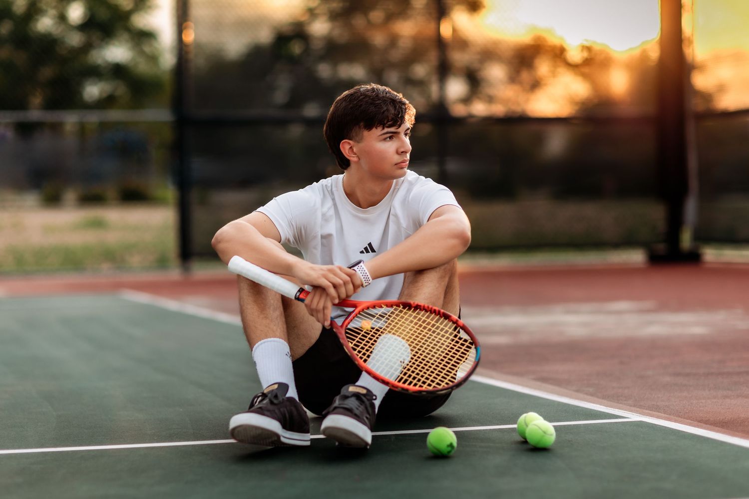 Person with tennis racket and balls sits on outdoor court during sunset session in Navarre Florida