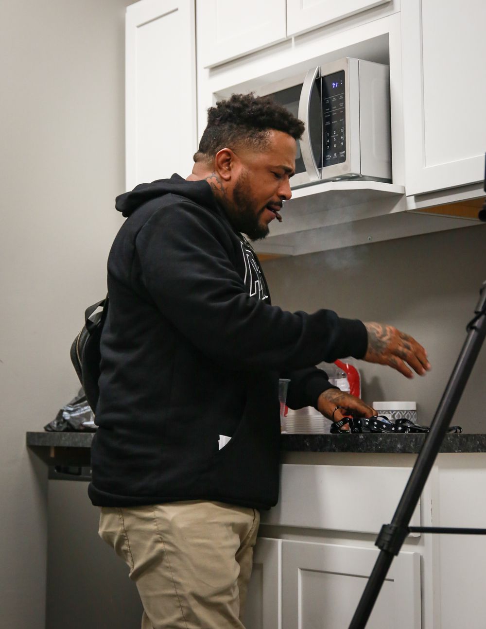 Someone wearing a black hoodie reaching for cooking controls on a stovetop while holding an electronic device in the kitchen.