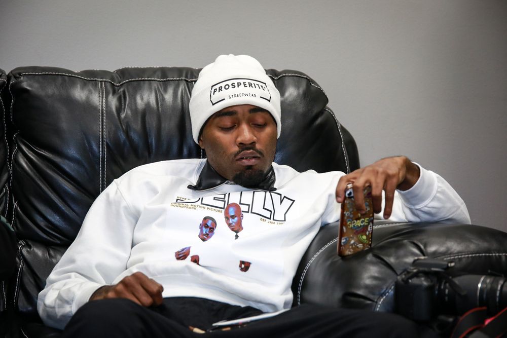 A person in a white beanie and sweatshirt relaxes on a black leather couch while holding a drink.