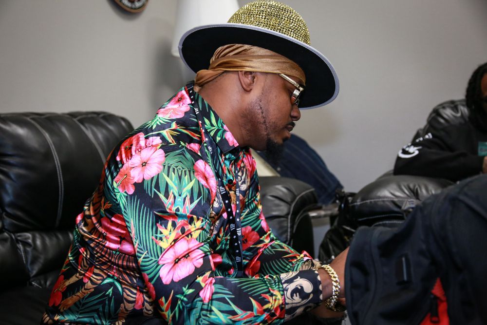 Someone wearing a colorful tropical print shirt and a hat sits on a black leather couch looking at their phone.