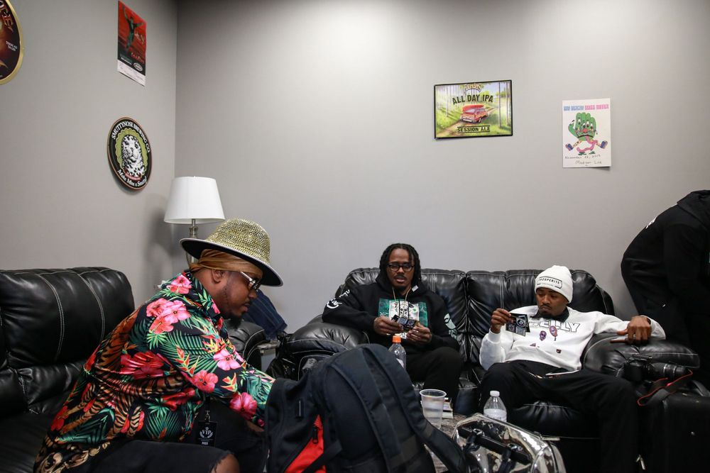 A group relaxes on black leather couches in a small room with pictures on gray walls and dim lighting.