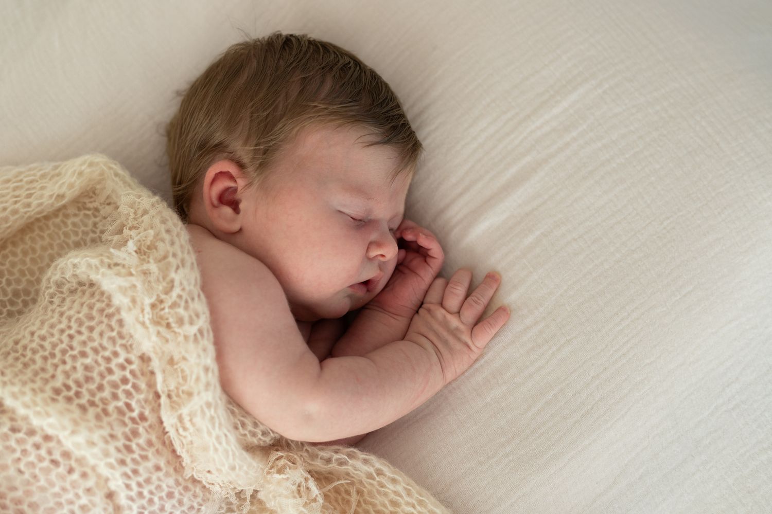 A sleeping newborn baby wrapped in cream-colored knit blanket on white textured background.