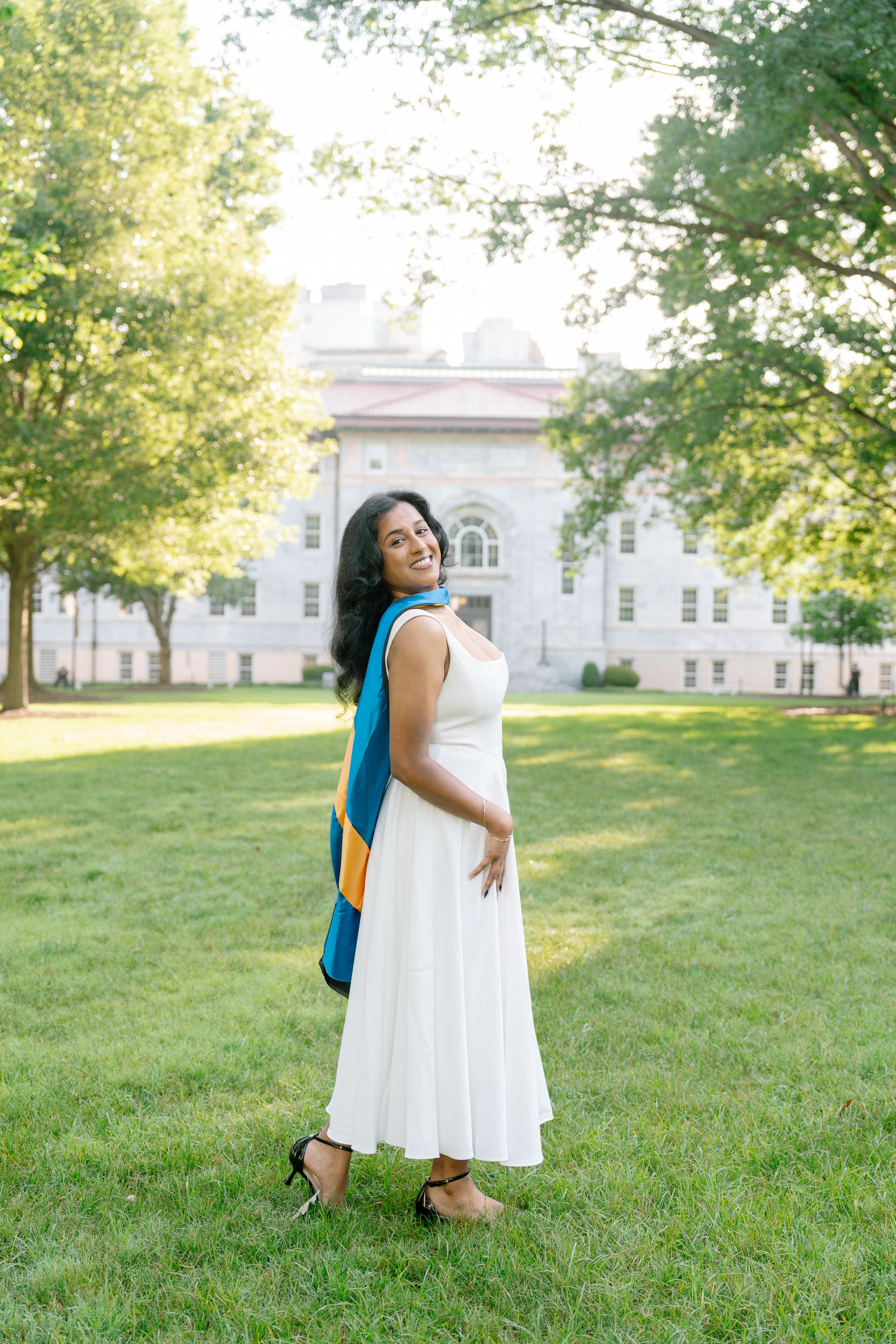 Woman in white dress and blue graduation hood smiling on green campus lawn near classic brick building.