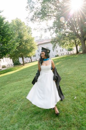 Smiling graduate in white dress and cap twirling on green campus lawn with sunlight streaming through trees.