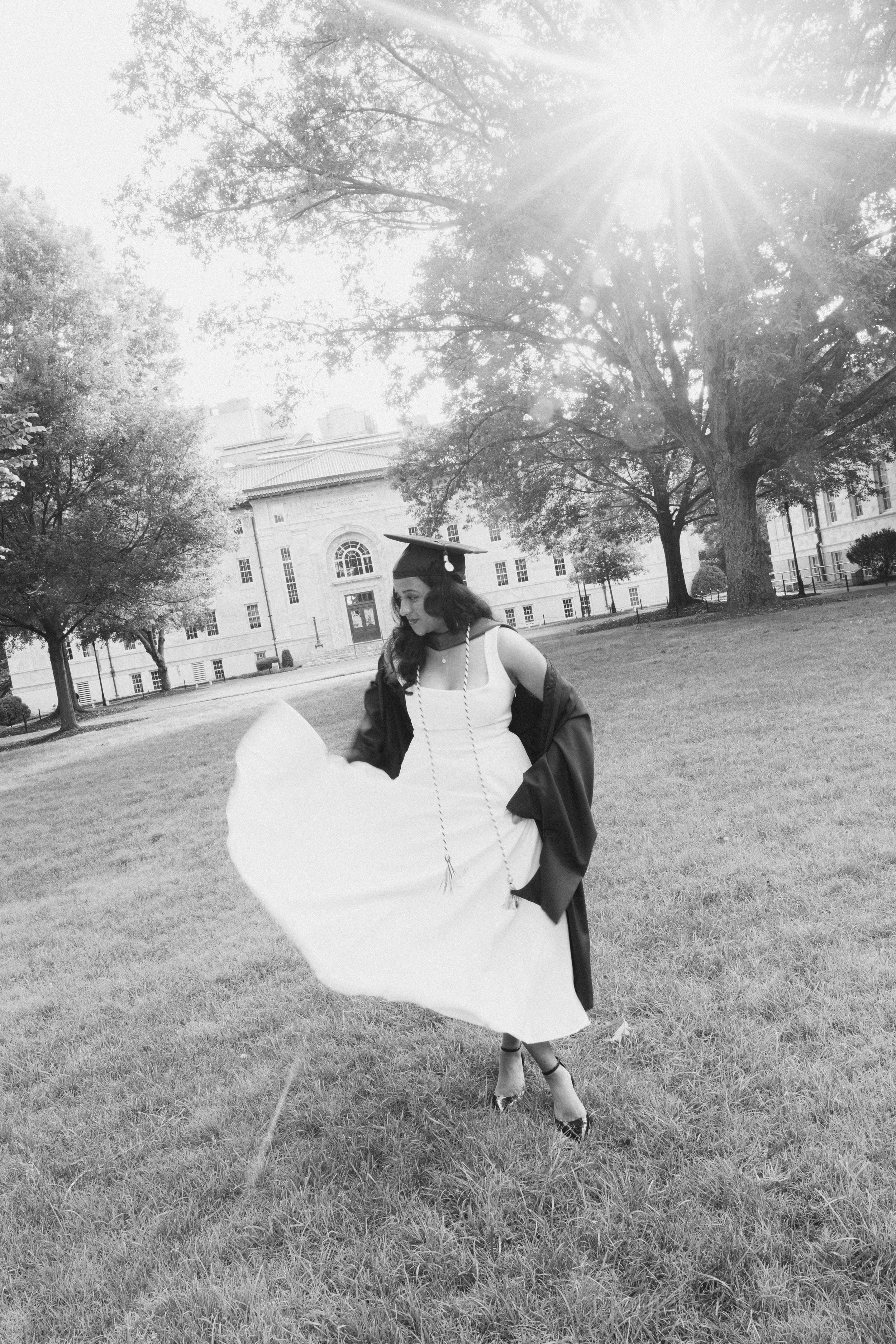 Black and white photo of a graduate in cap and gown twirling on a sunny campus lawn.
