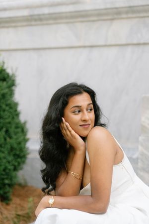 A young woman in a white dress with gold bracelets poses gracefully outdoors near green shrubs and a marble wall.