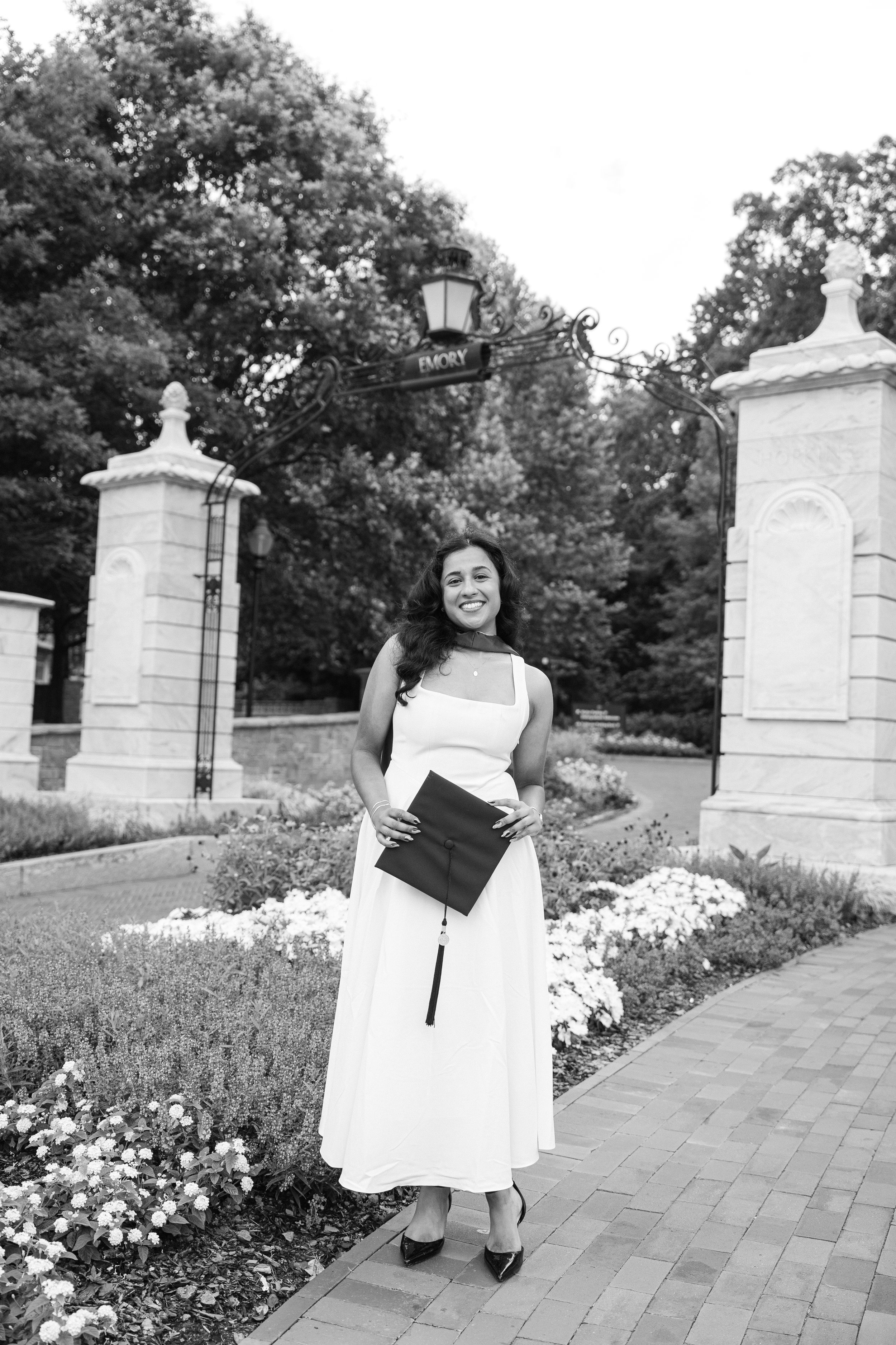 Graduate in white dress holding cap smiles at Emory University entrance gate in black and white photo.
