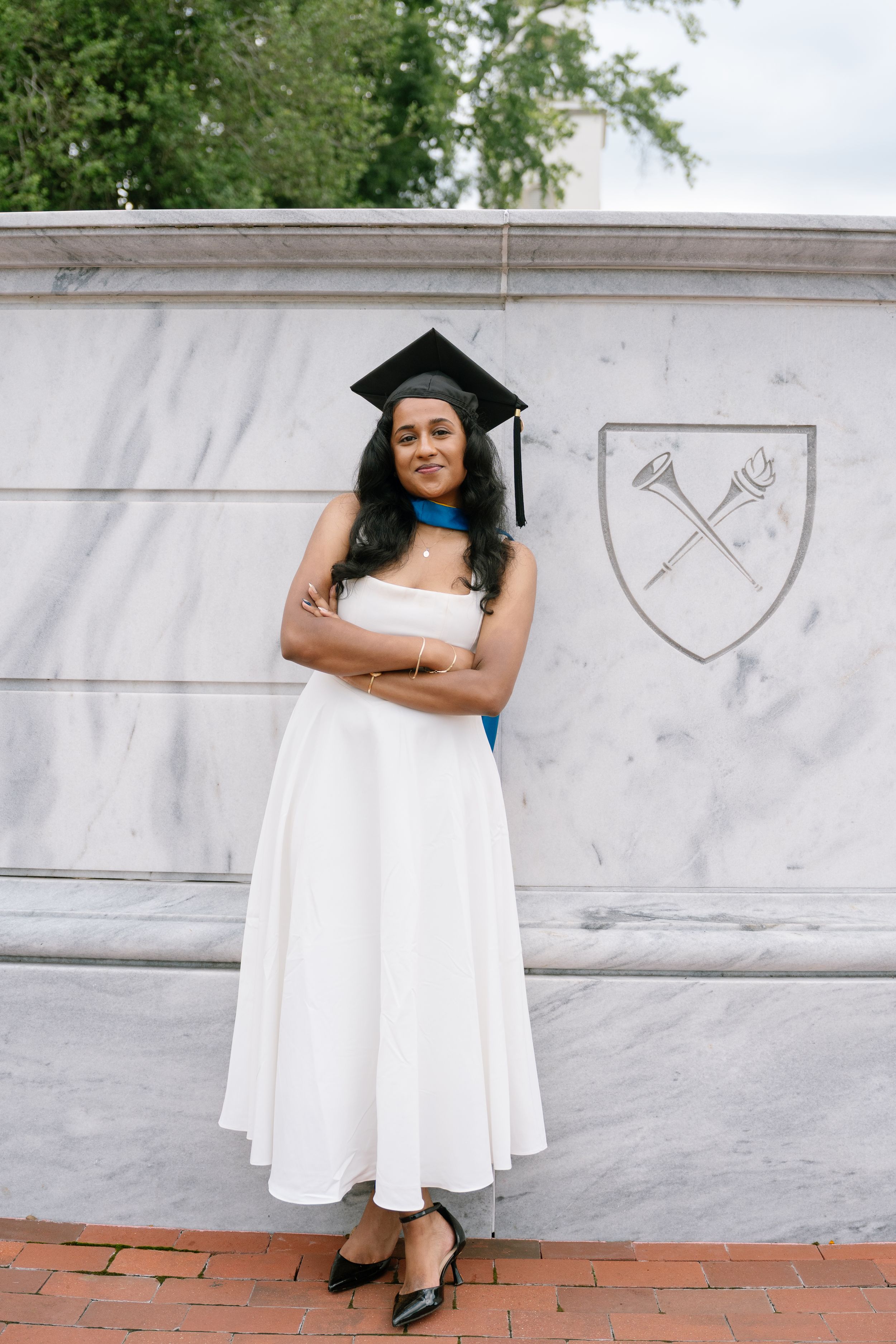 A young woman in a white dress and graduation cap poses confidently with arms crossed in front of a university emblem.