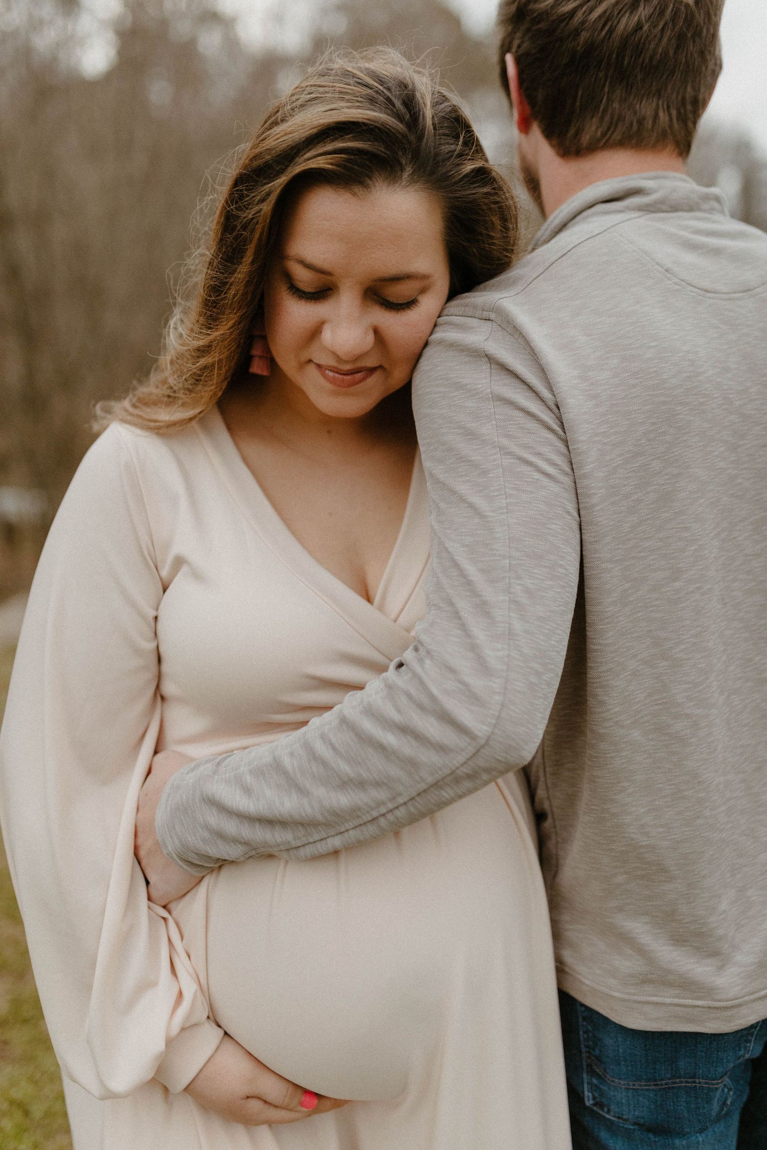 Intimate maternity photo of couple embracing outdoors, woman wearing cream wrap dress.