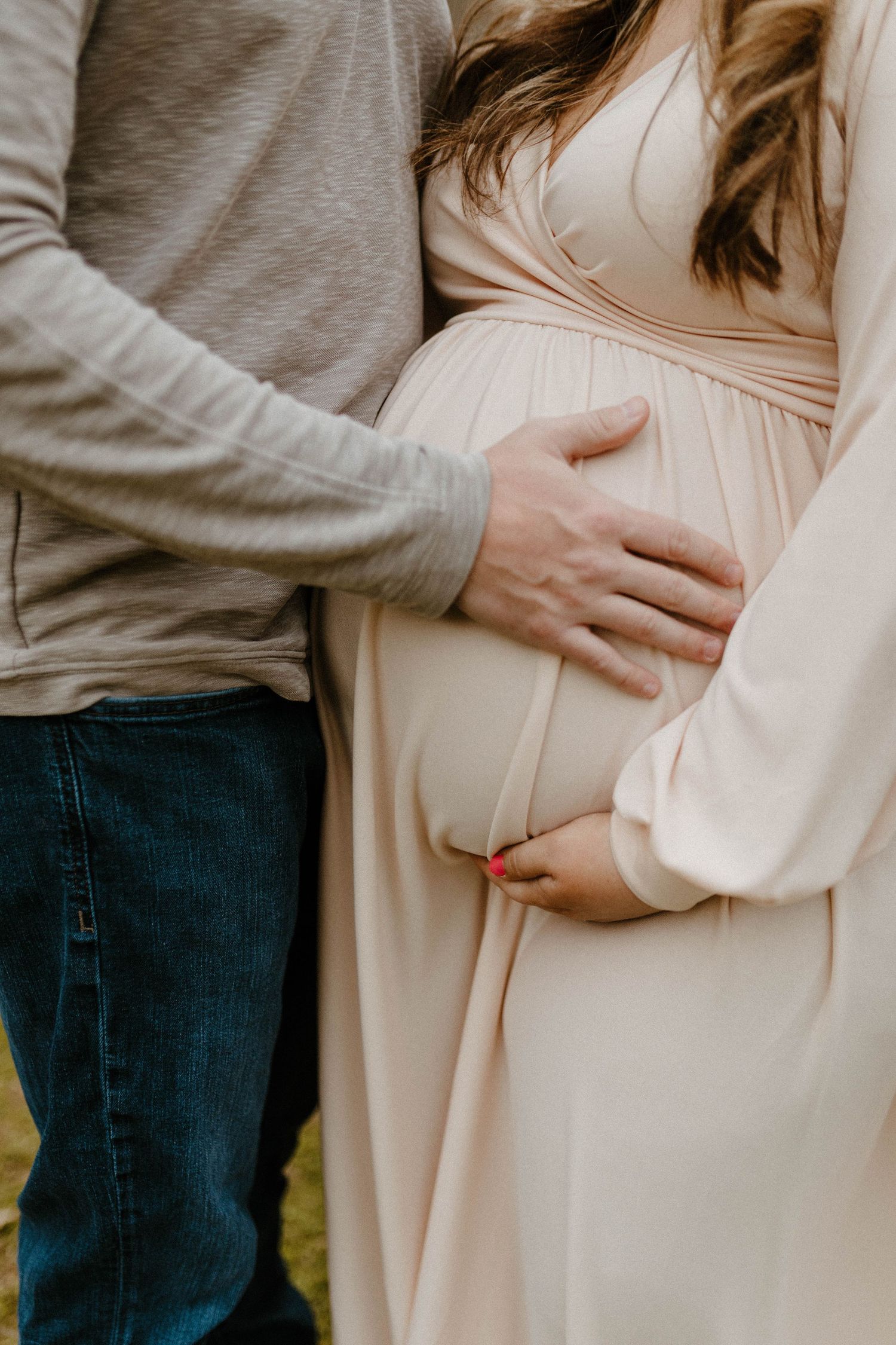Close-up of couple's hands resting on pregnant belly in cream-colored maternity dress.