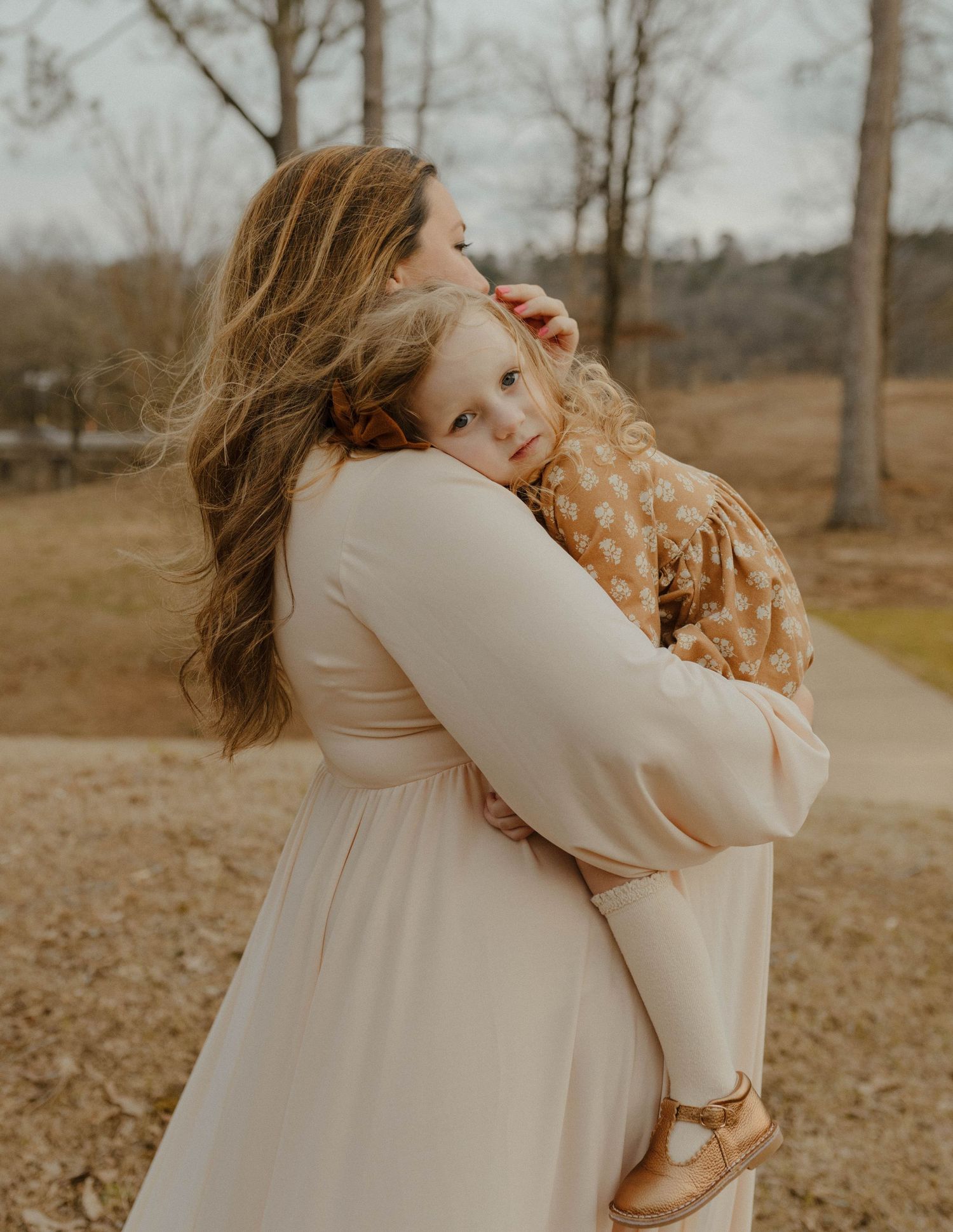 Mother in cream dress embraces small child wearing floral pattern dress in outdoor autumn setting.