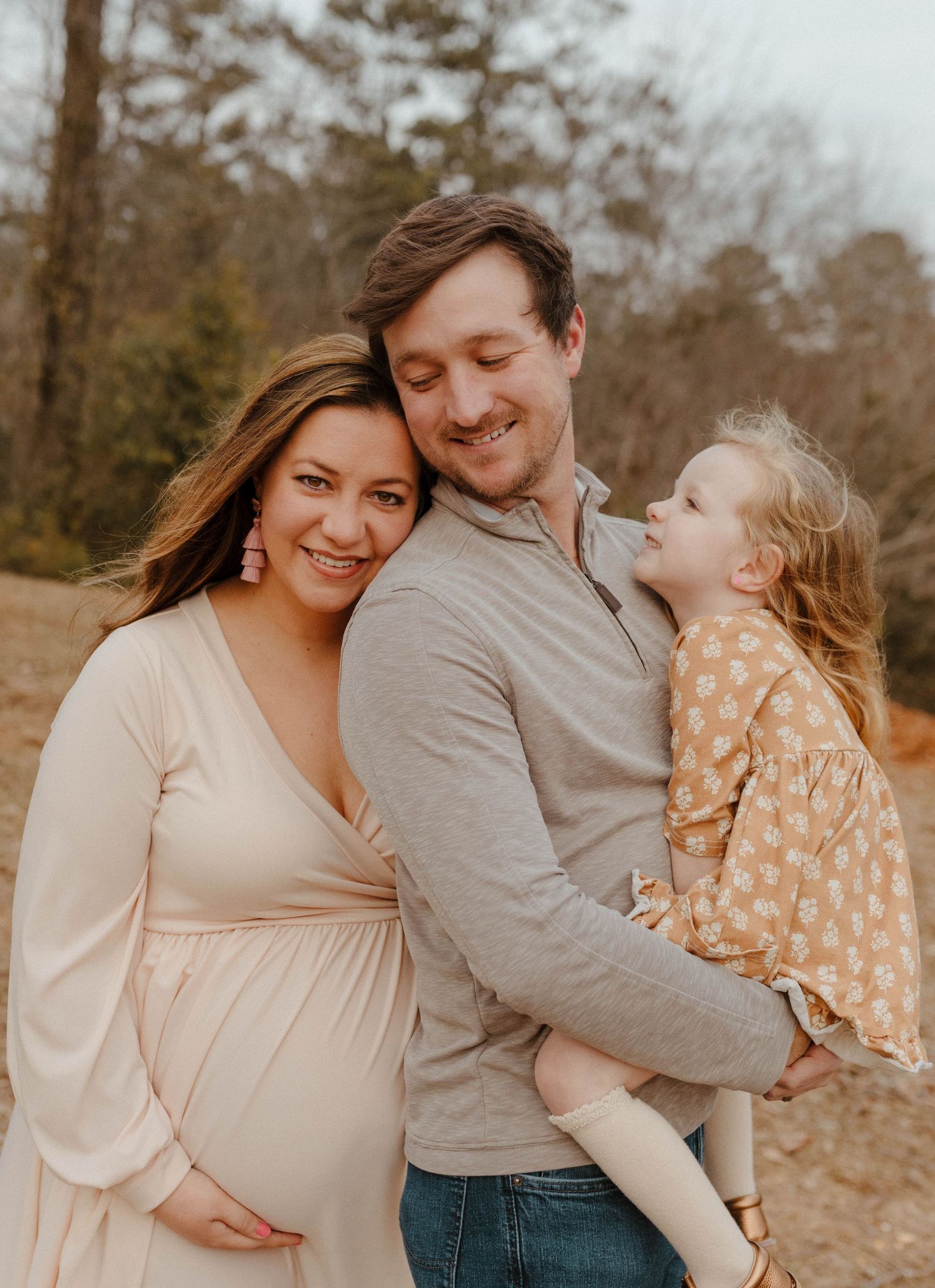 A family portrait in warm tones showing a pregnant woman in cream dress with a toddler in floral dress and partner in grey.