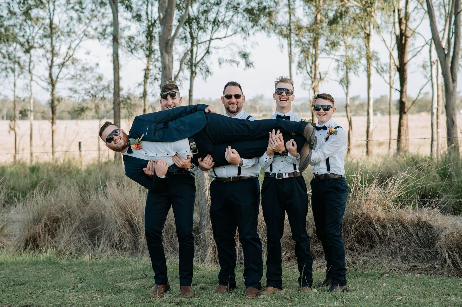Groomsmen in suspenders playfully carrying someone horizontal for a fun wedding photo.