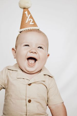 An excited baby wearing a tan collared shirt and paper party hat laughs with pure joy.