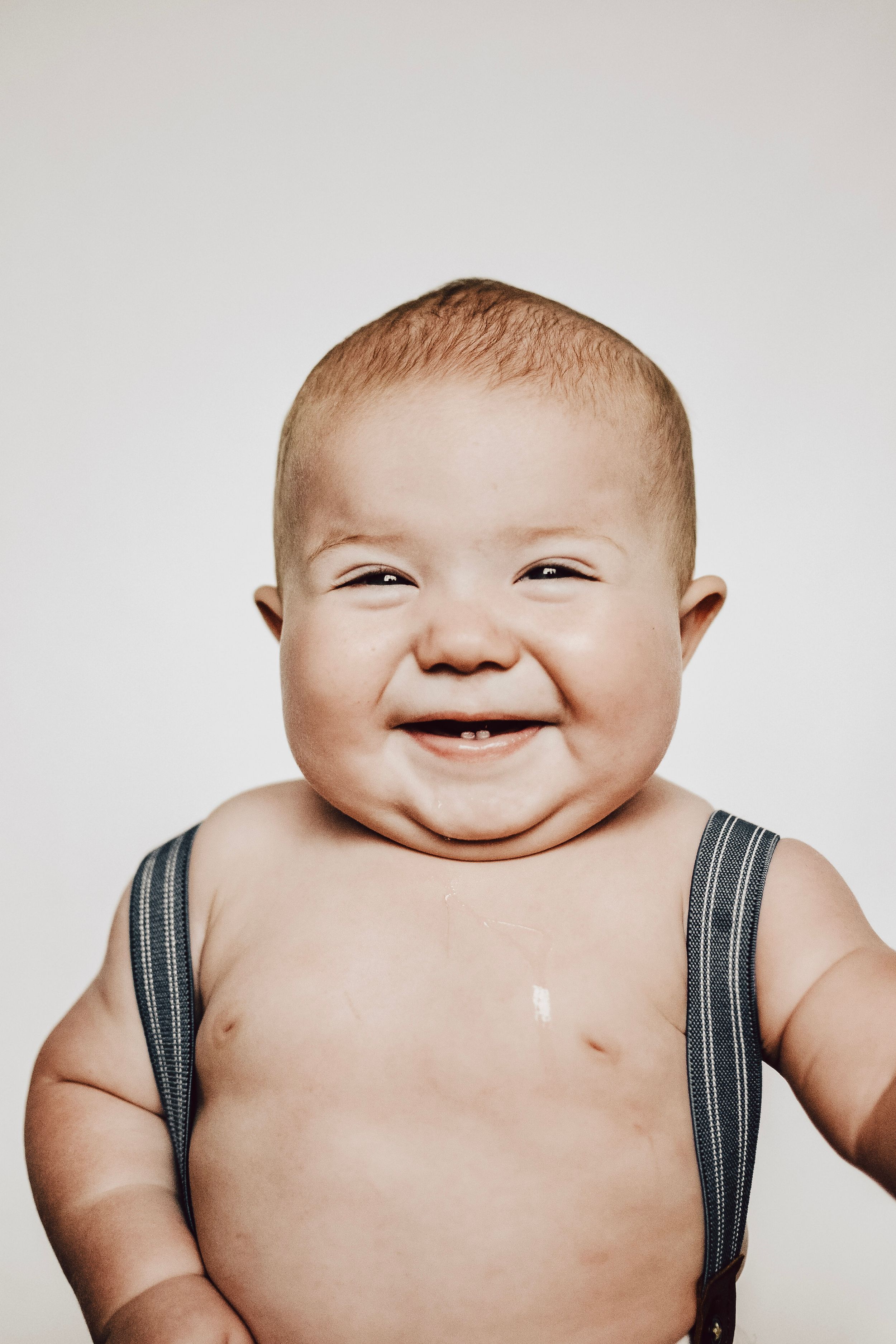 Portrait of a laughing baby wearing black straps against light background.