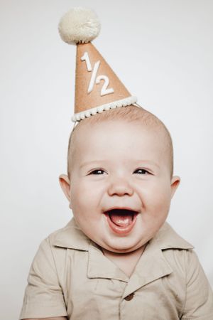 An enthusiastic baby wearing a tan shirt and party hat displays pure joy and laughter.