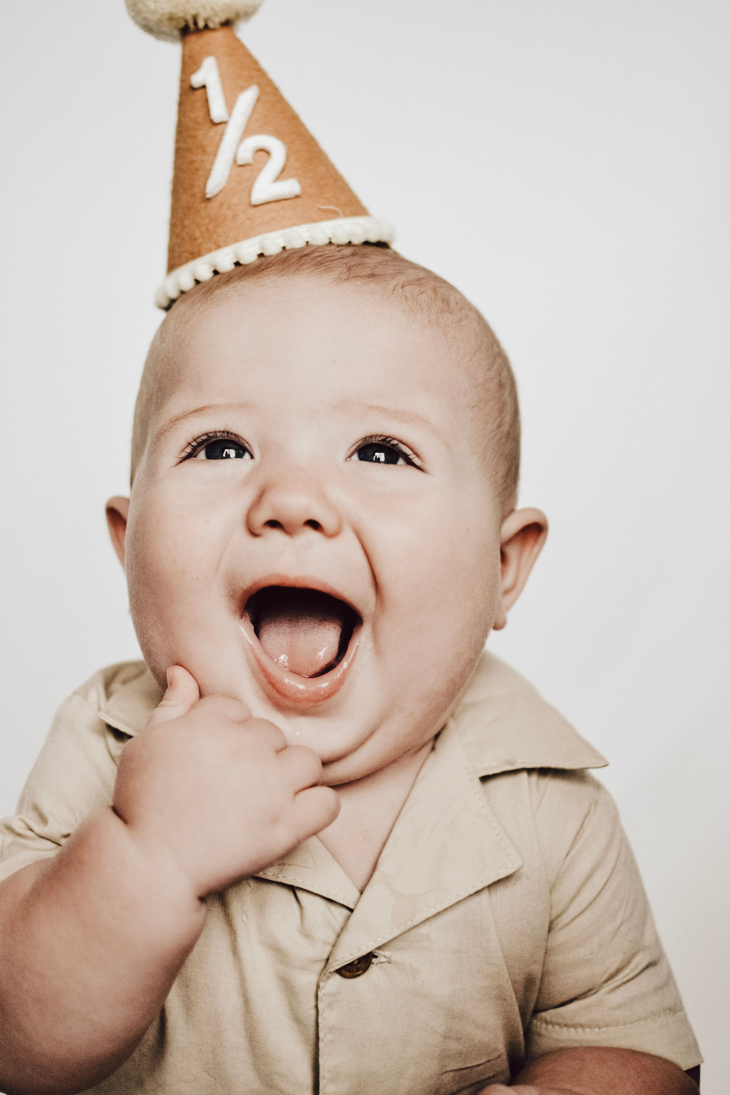 A cheerful baby in a beige shirt wears a '1/2' birthday party hat while giggling.
