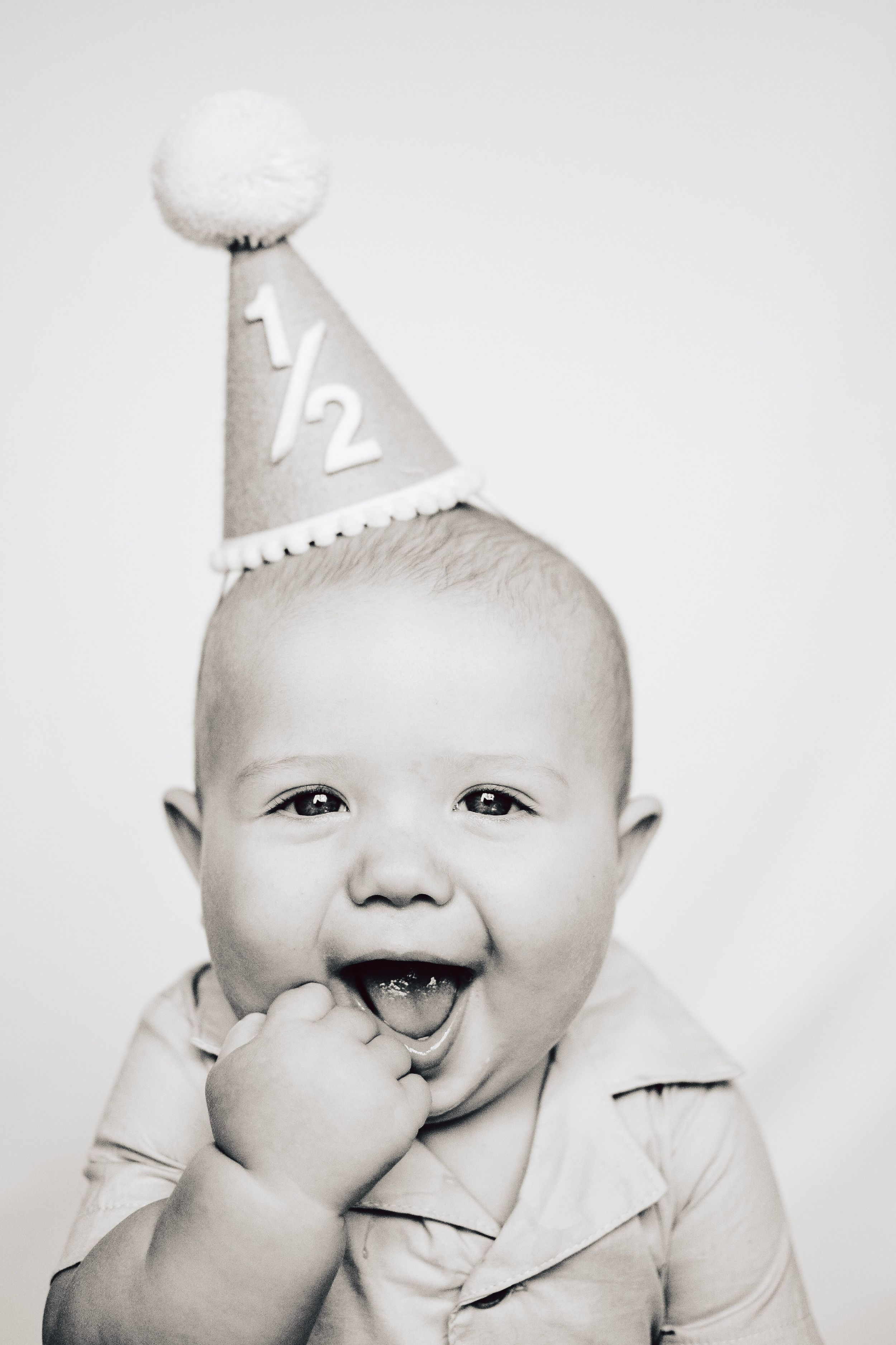 A delighted baby in a light colored shirt giggles while wearing a celebratory half-birthday hat.