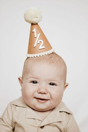 A happy baby in a light colored outfit smiles while wearing a '1/2' birthday celebration hat.