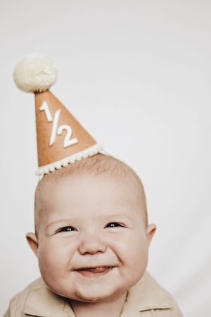 A cheerful baby in a beige shirt shows a big grin while wearing a '1/2' birthday hat.