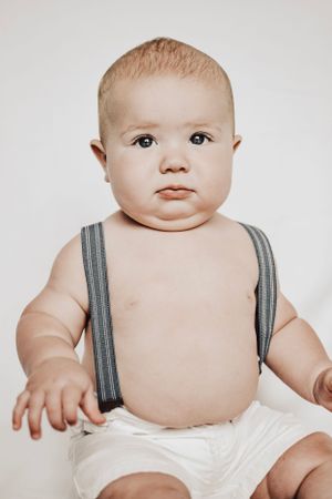 A baby in striped suspenders and white bottoms sits against a light background.