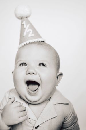 A delighted baby wearing a half birthday party hat and collared shirt laughs with excitement.