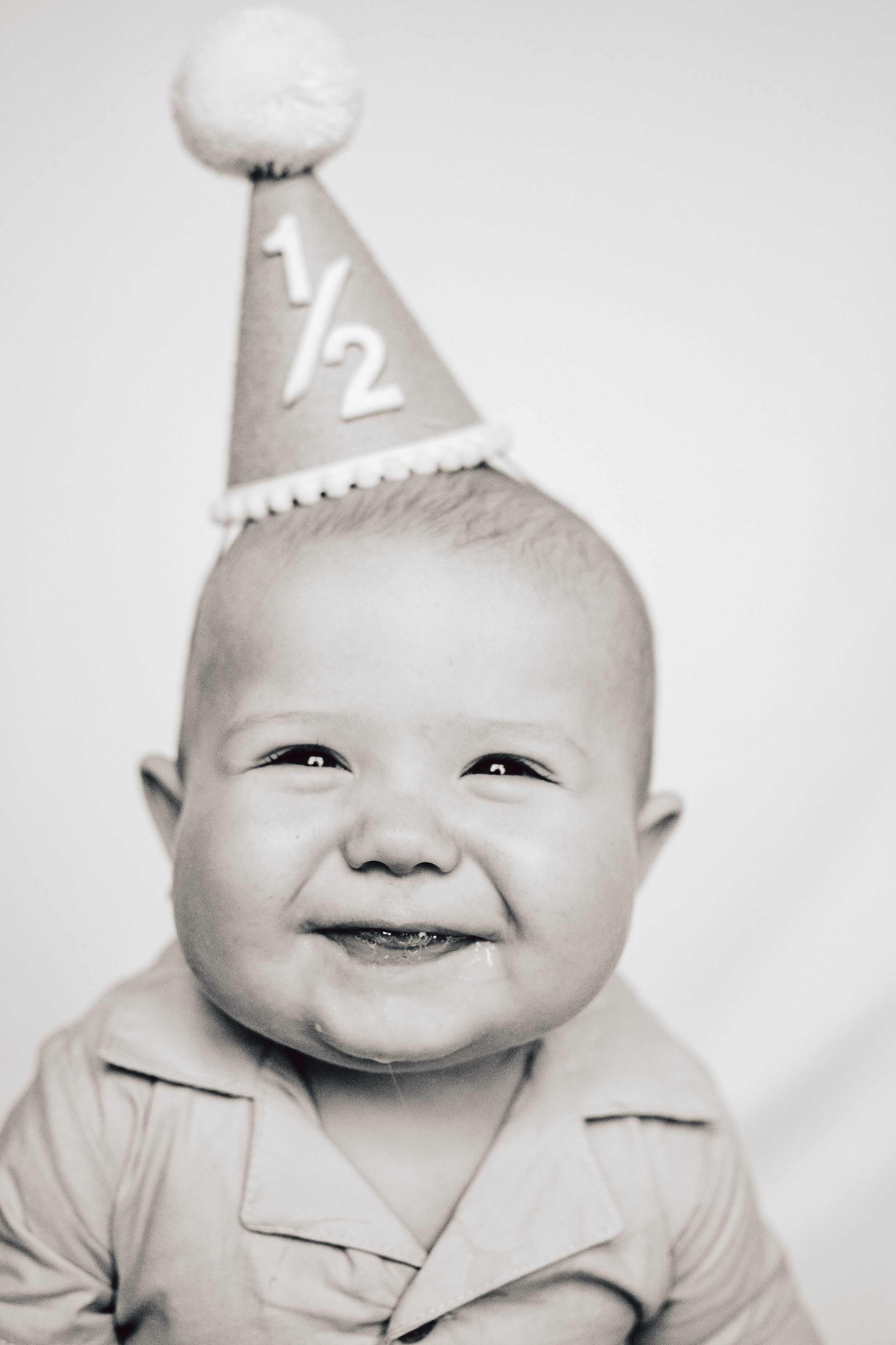 A content baby wearing a half-birthday party hat and collared shirt gives a sweet smile.