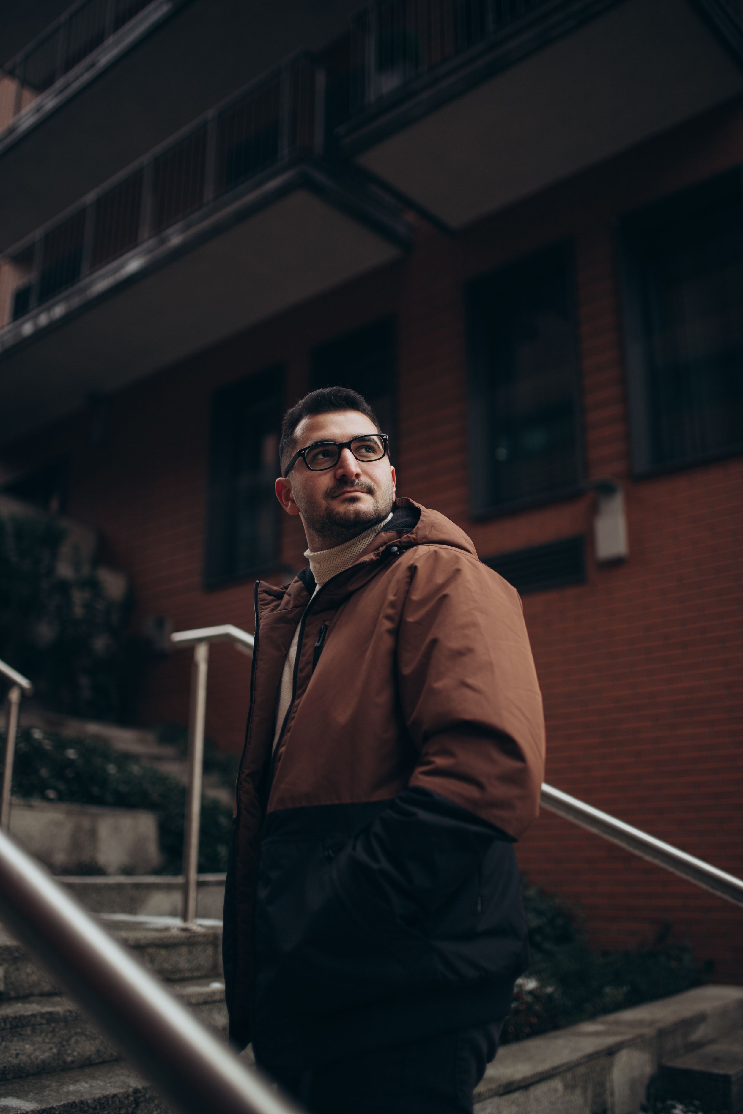 Low angle urban portrait featuring brown jacket and glasses against brick building architecture.