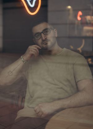 Atmospheric portrait through window glass showing contemplative pose in beige t-shirt and glasses.