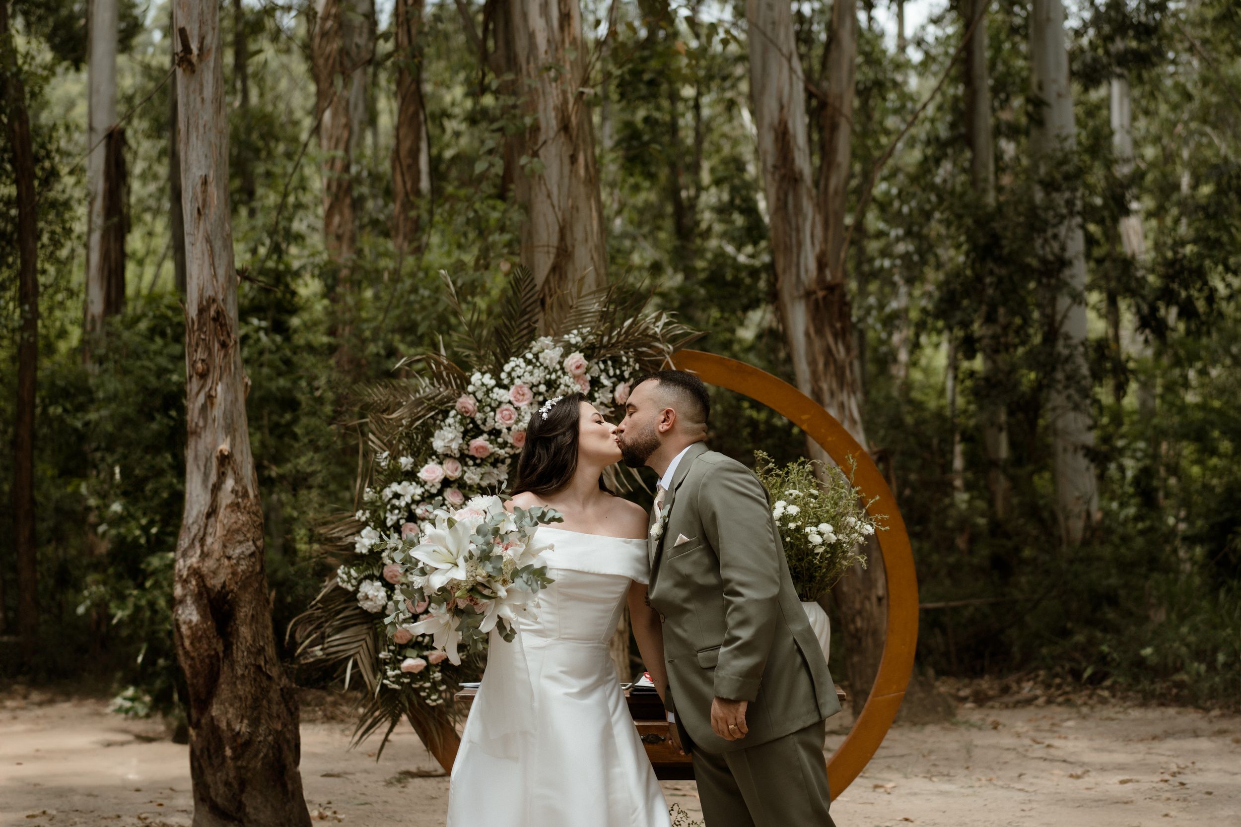 Couple shares intimate moment under circular floral arch in eucalyptus forest setting.