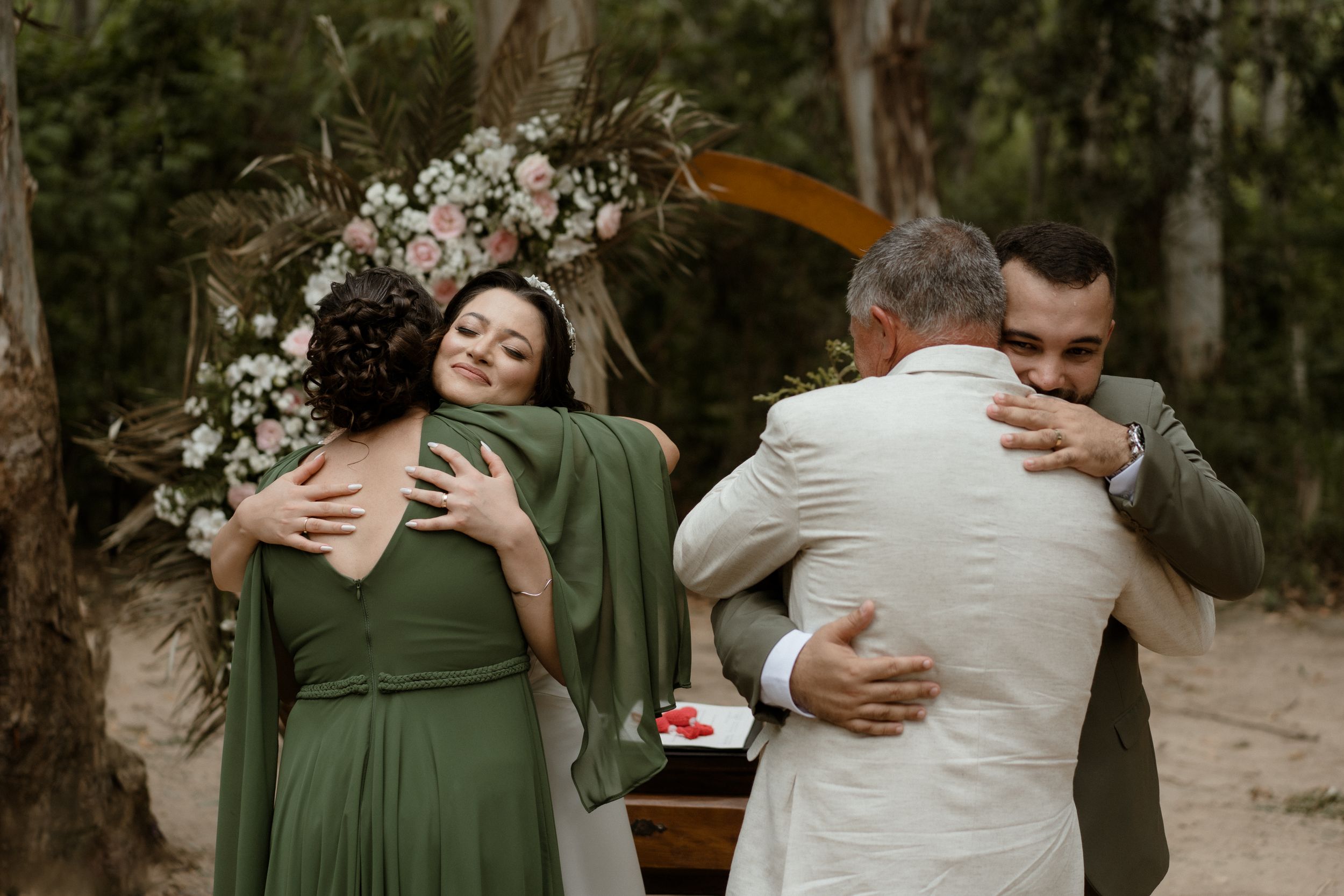 Wedding party shares emotional hugs during outdoor forest ceremony with floral arch and olive green attire.
