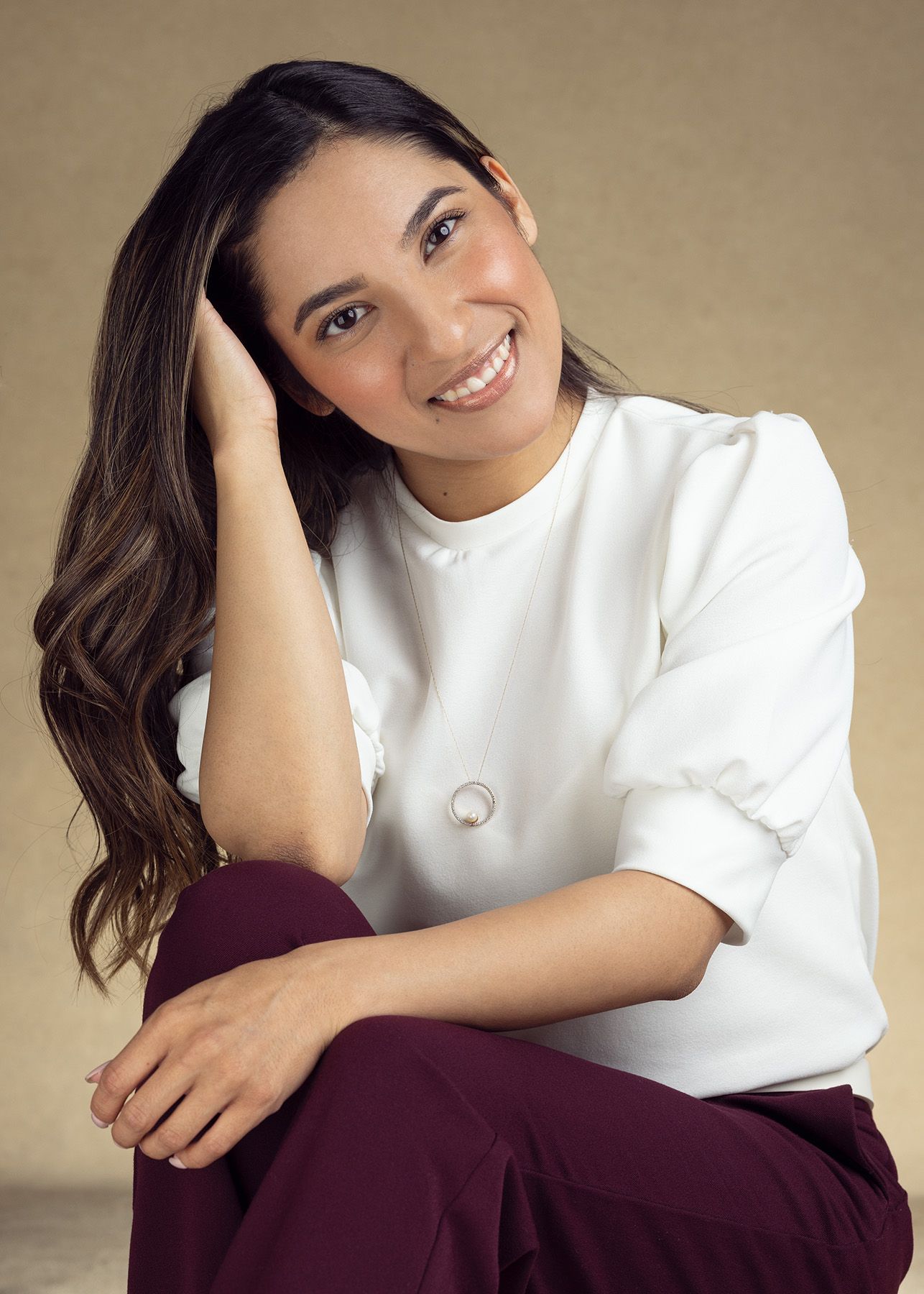 A figure in a casual white sweater and burgundy pants sits casually against a neutral backdrop with long wavy dark hair. photography