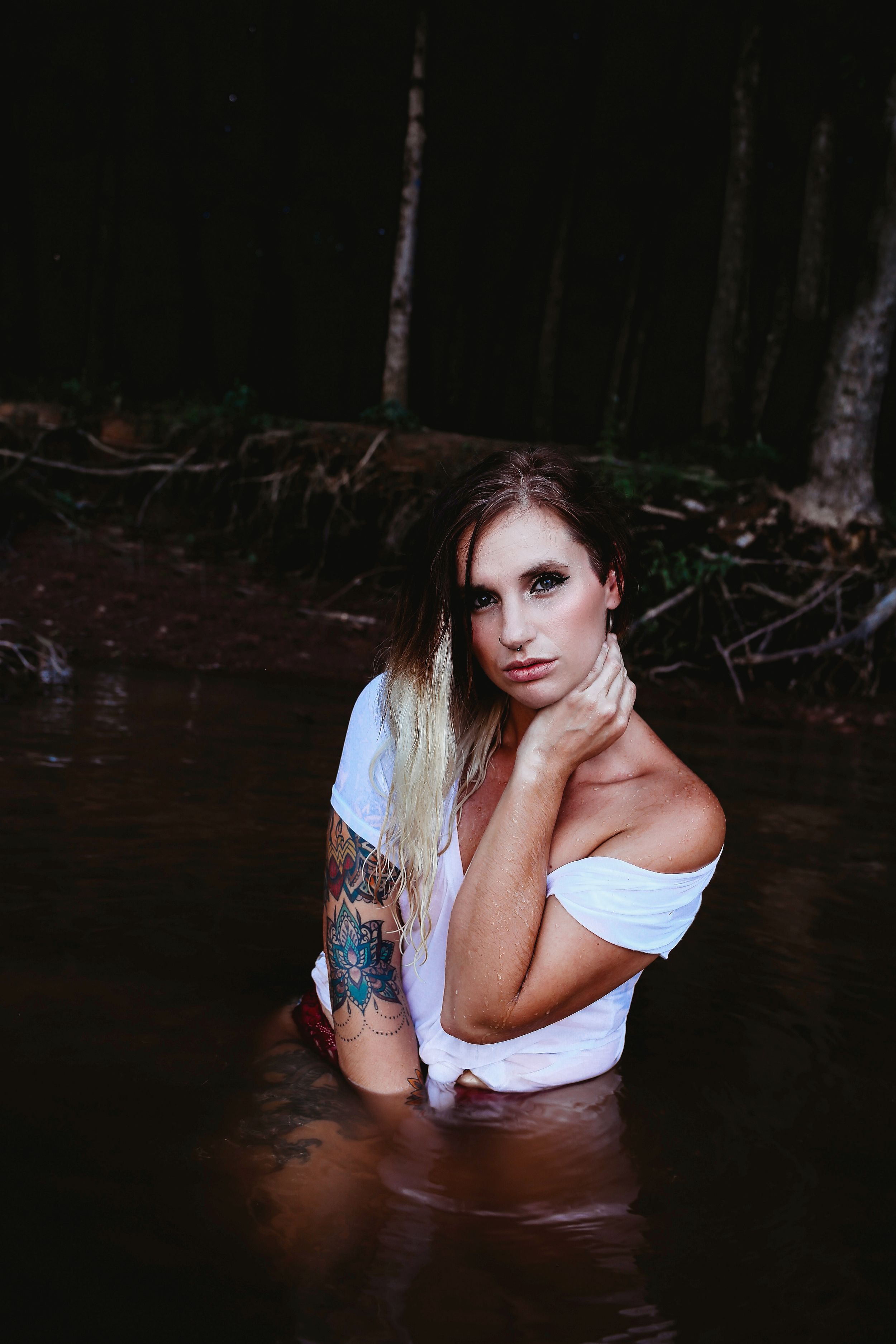 Artistic water portrait with white top and tattoos in dark forest setting.