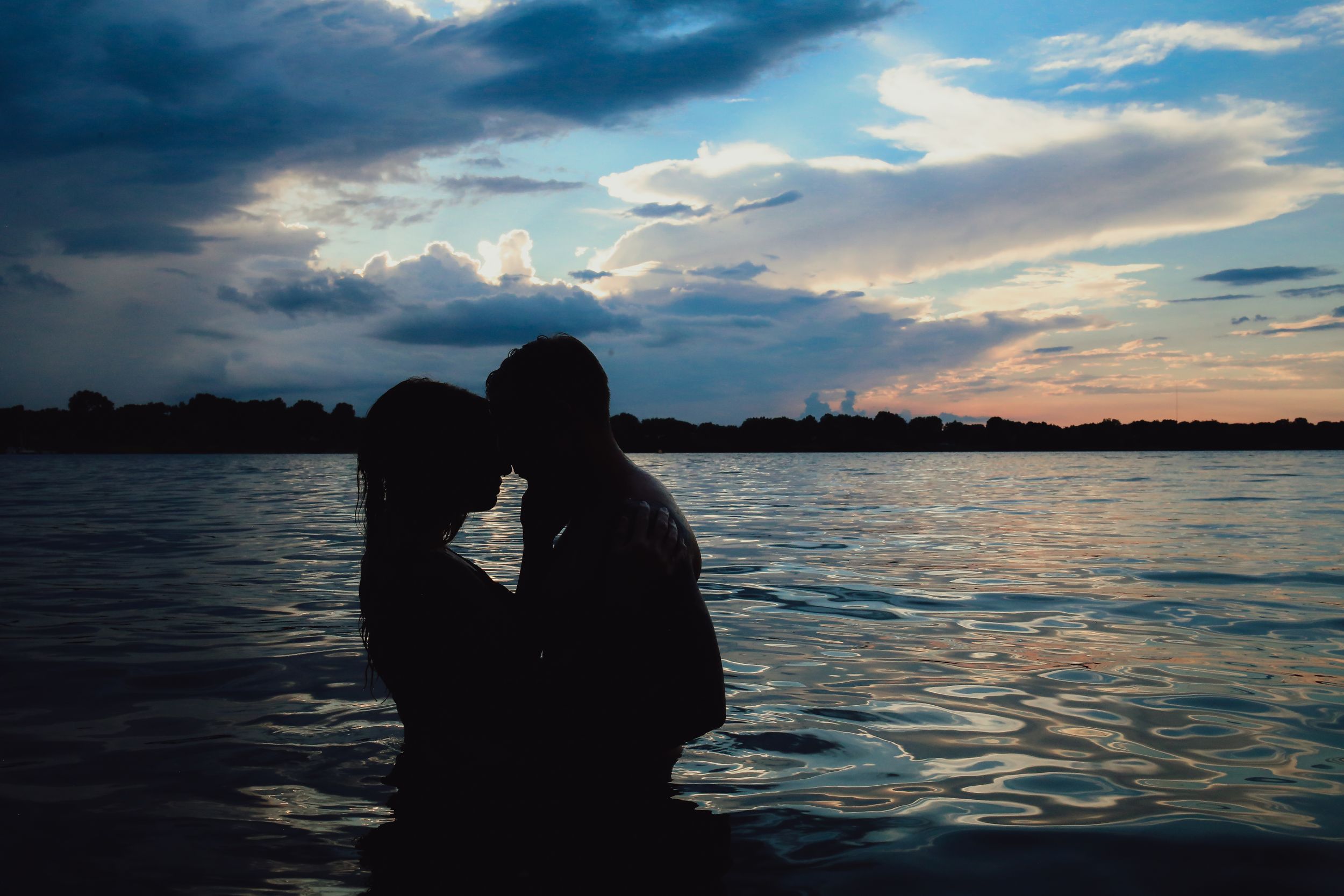 Silhouetted figures create romantic moment against dramatic sunset sky over water.
