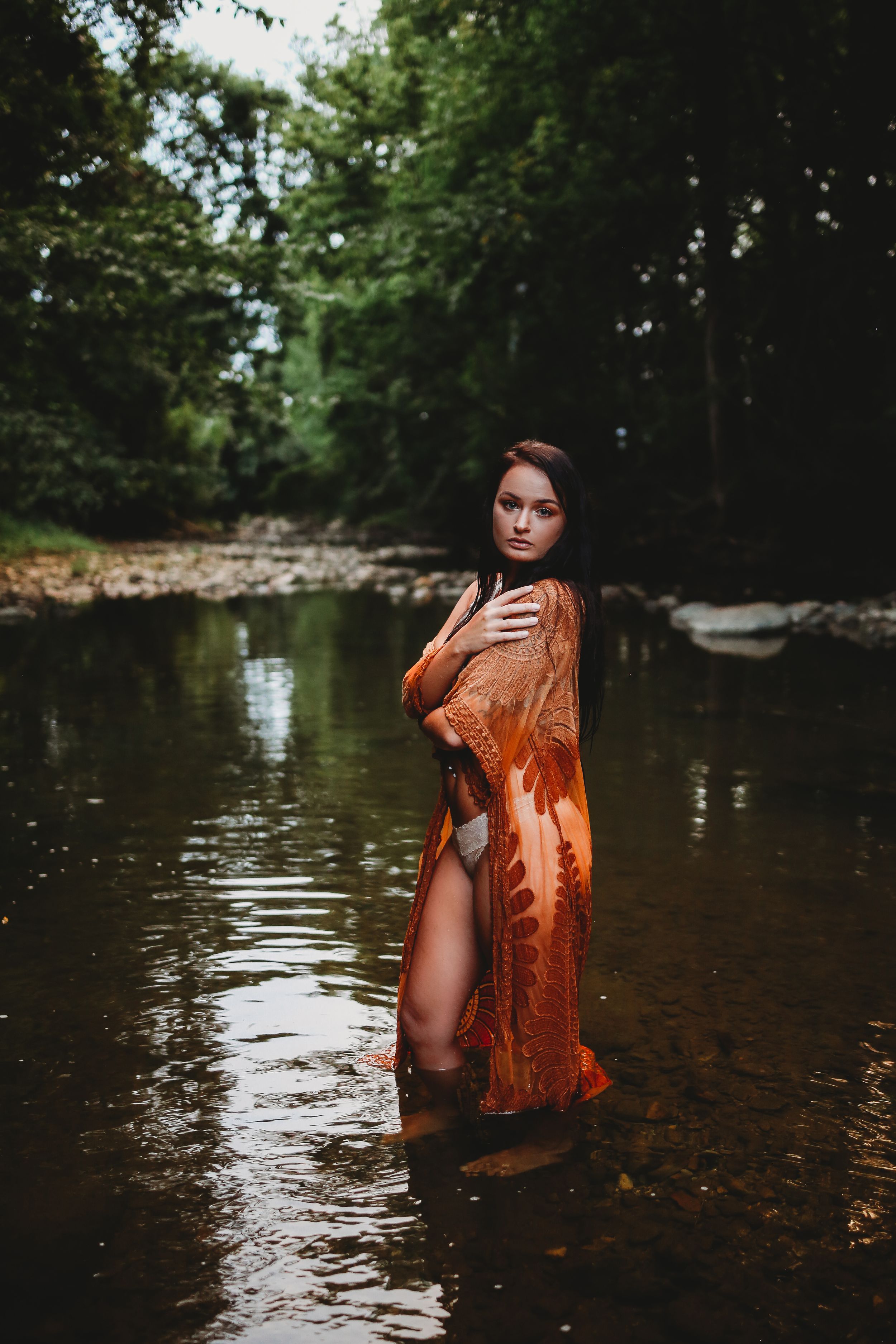 Artistic portrait in shallow water wearing orange lace dress among trees and natural scenery.