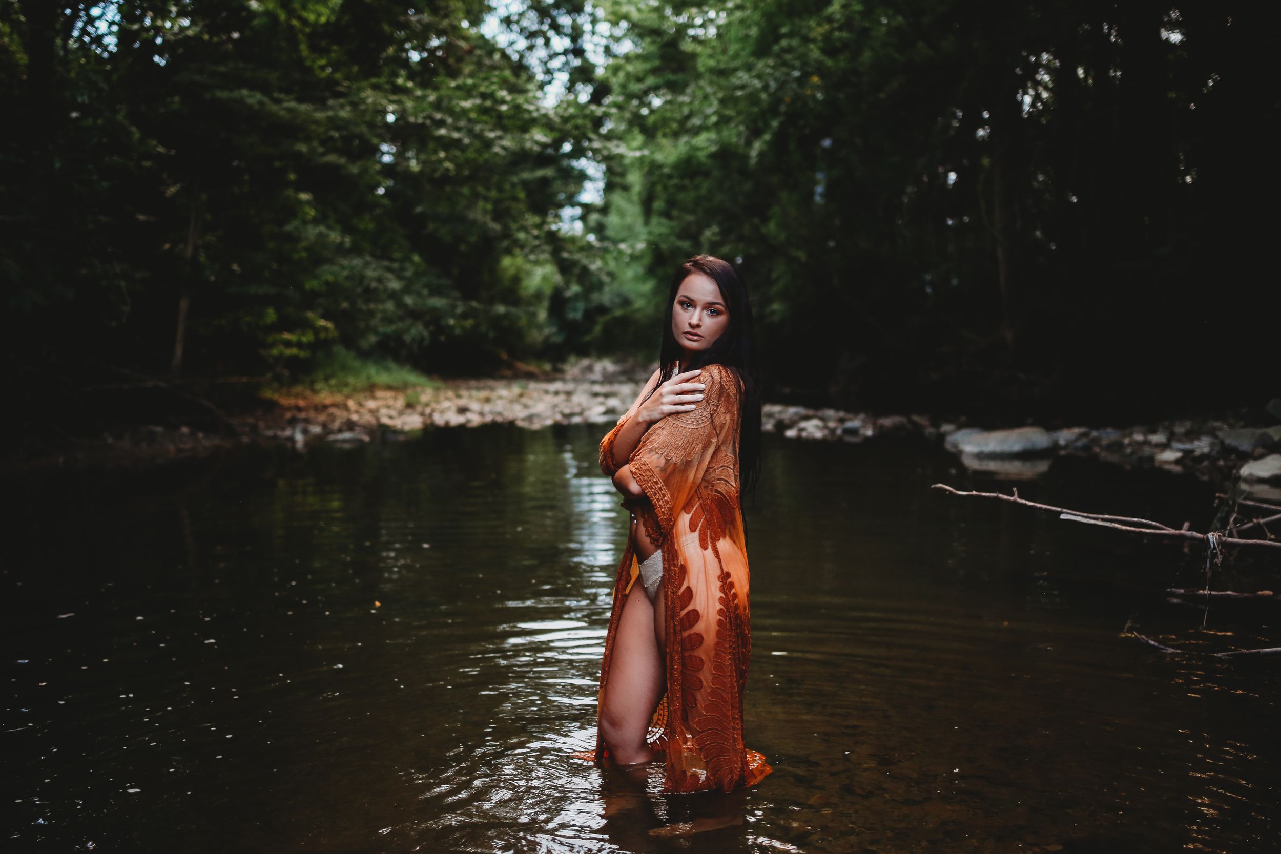 Model in orange dress photographed in shallow woodland stream with natural lighting.