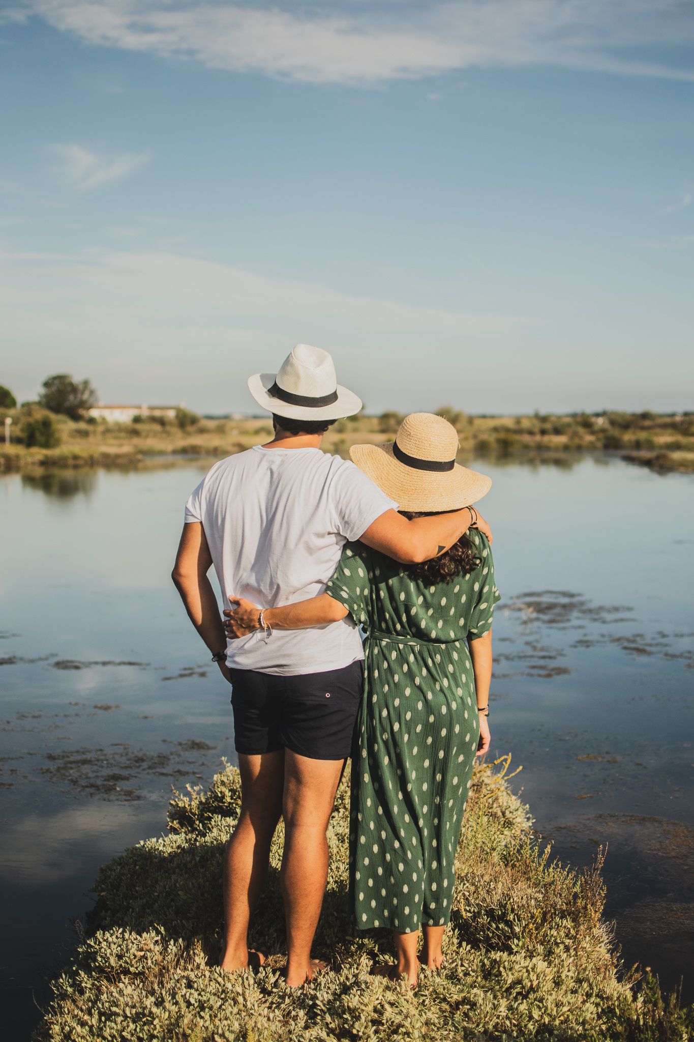 Couple in summer attire enjoying sunset view by lakeside wearing matching straw hats.
