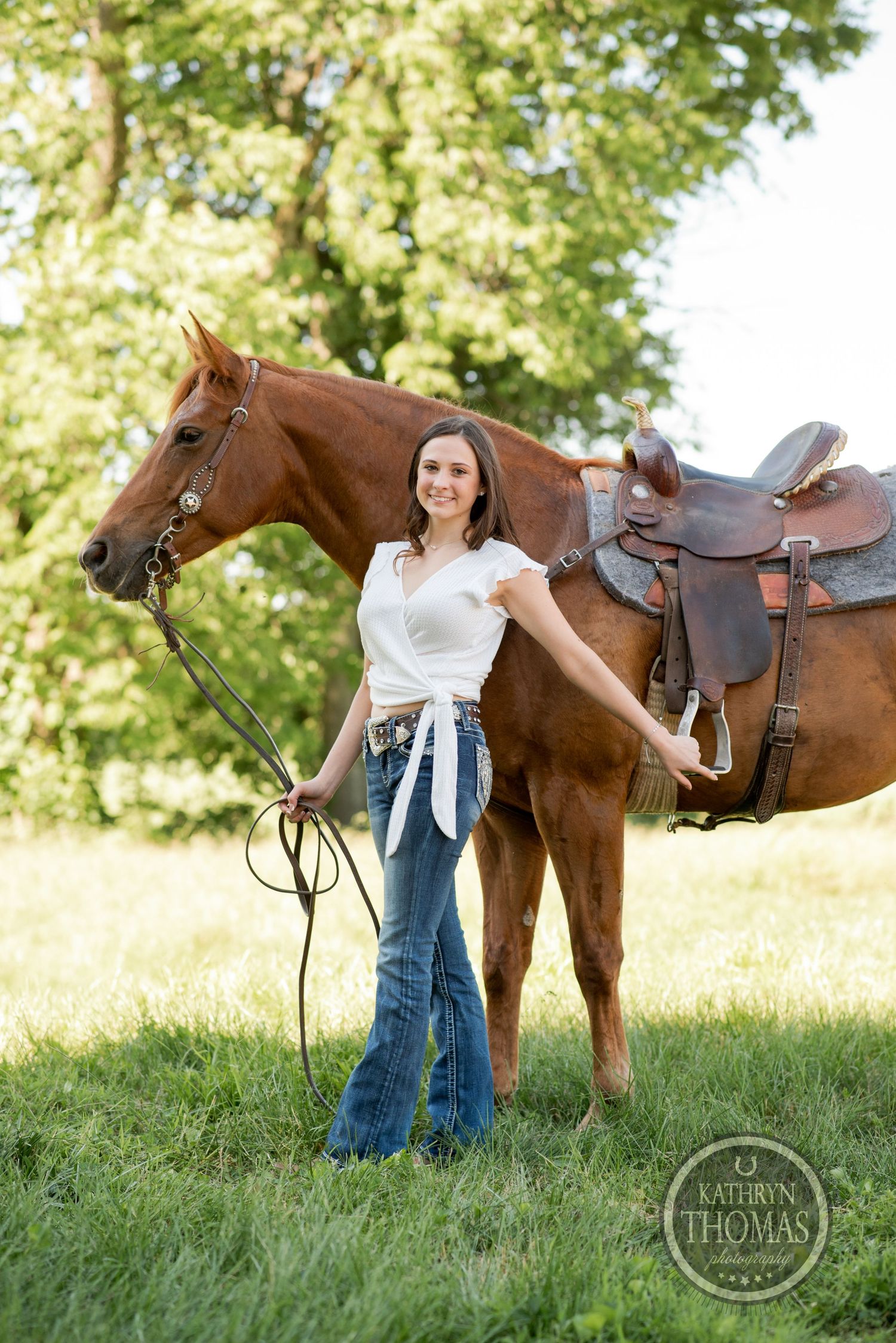 A woman in jeans and white top stands holding the reins of a saddled brown horse in a green field.