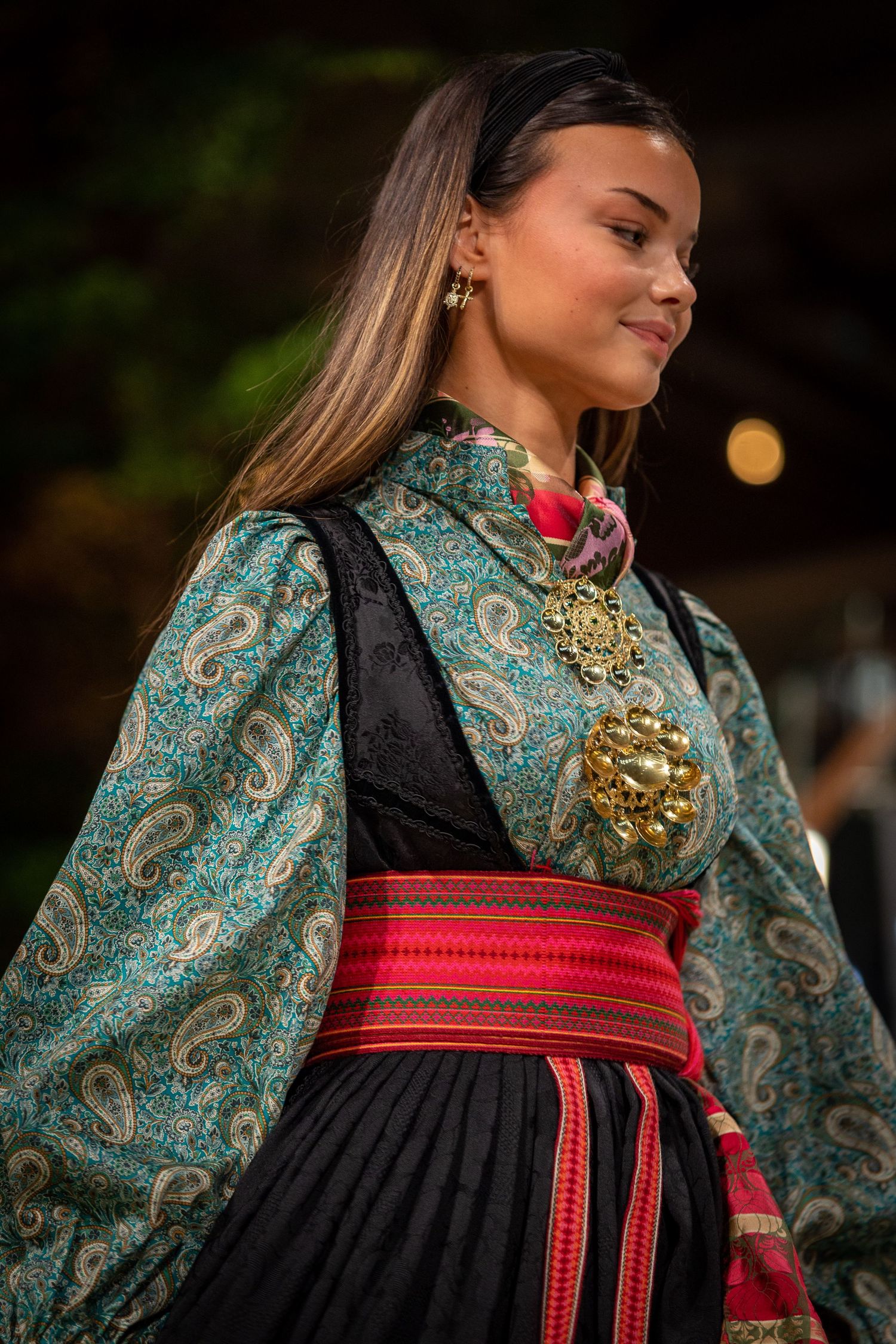 Young woman in traditional Norwegian bunad costume with paisley blouse, black vest, red sash, and gold brooch.