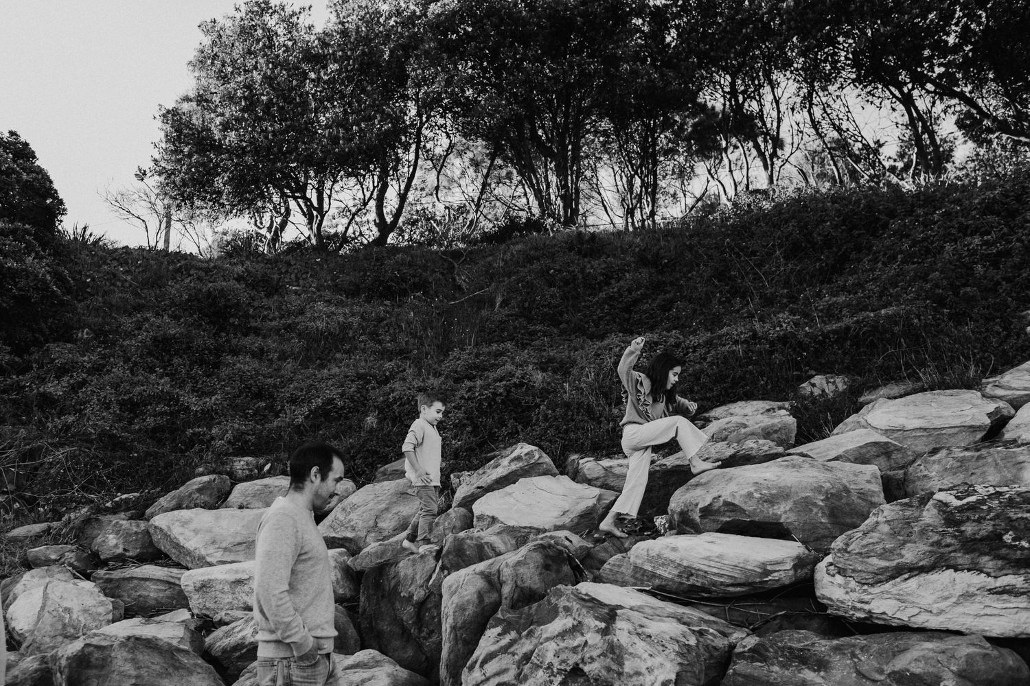 Black and white photo of family climbing over large coastal rocks with trees in background.