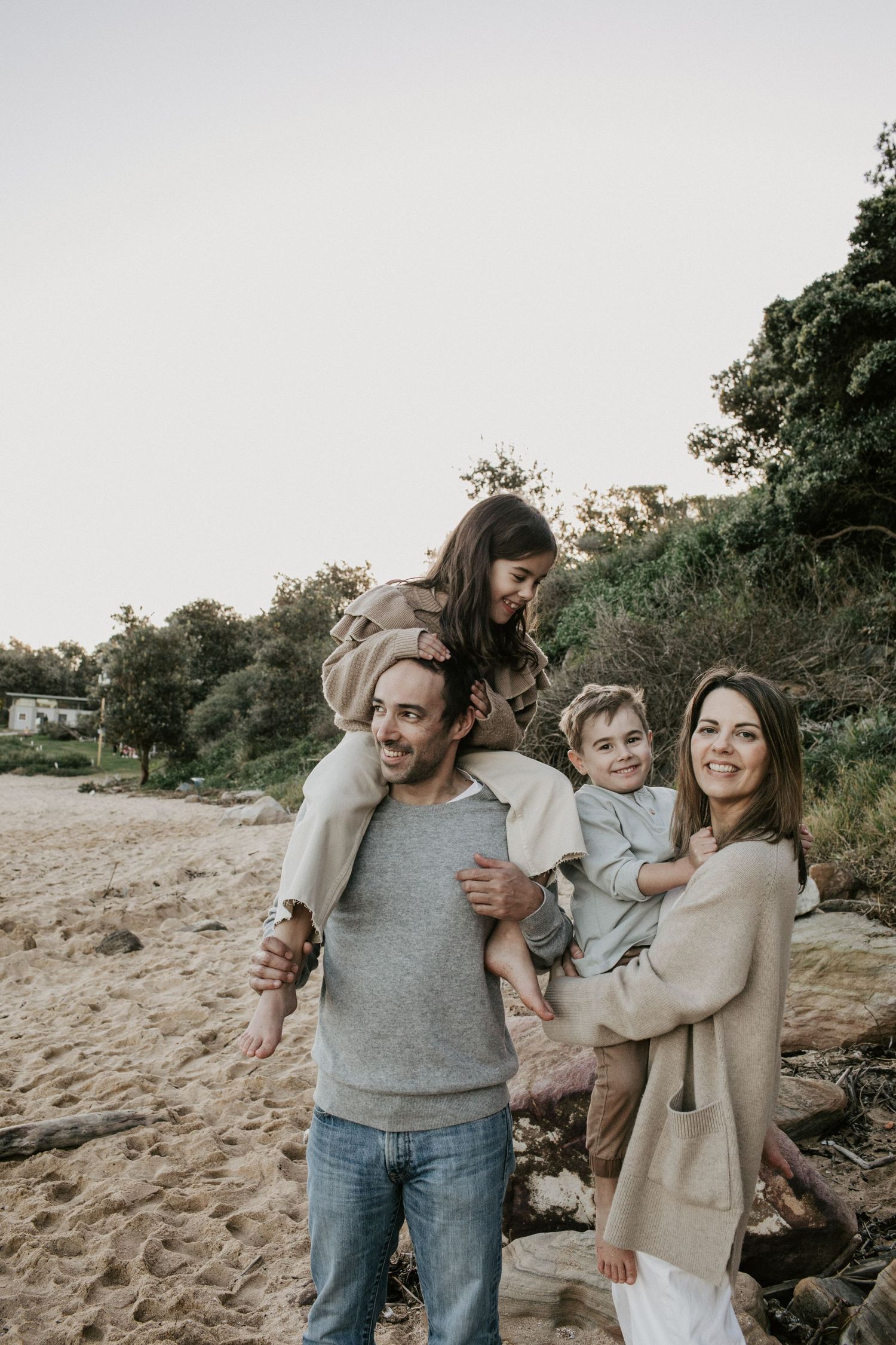 Happy family of four on a beach with dad carrying daughter on shoulders and mom holding son.